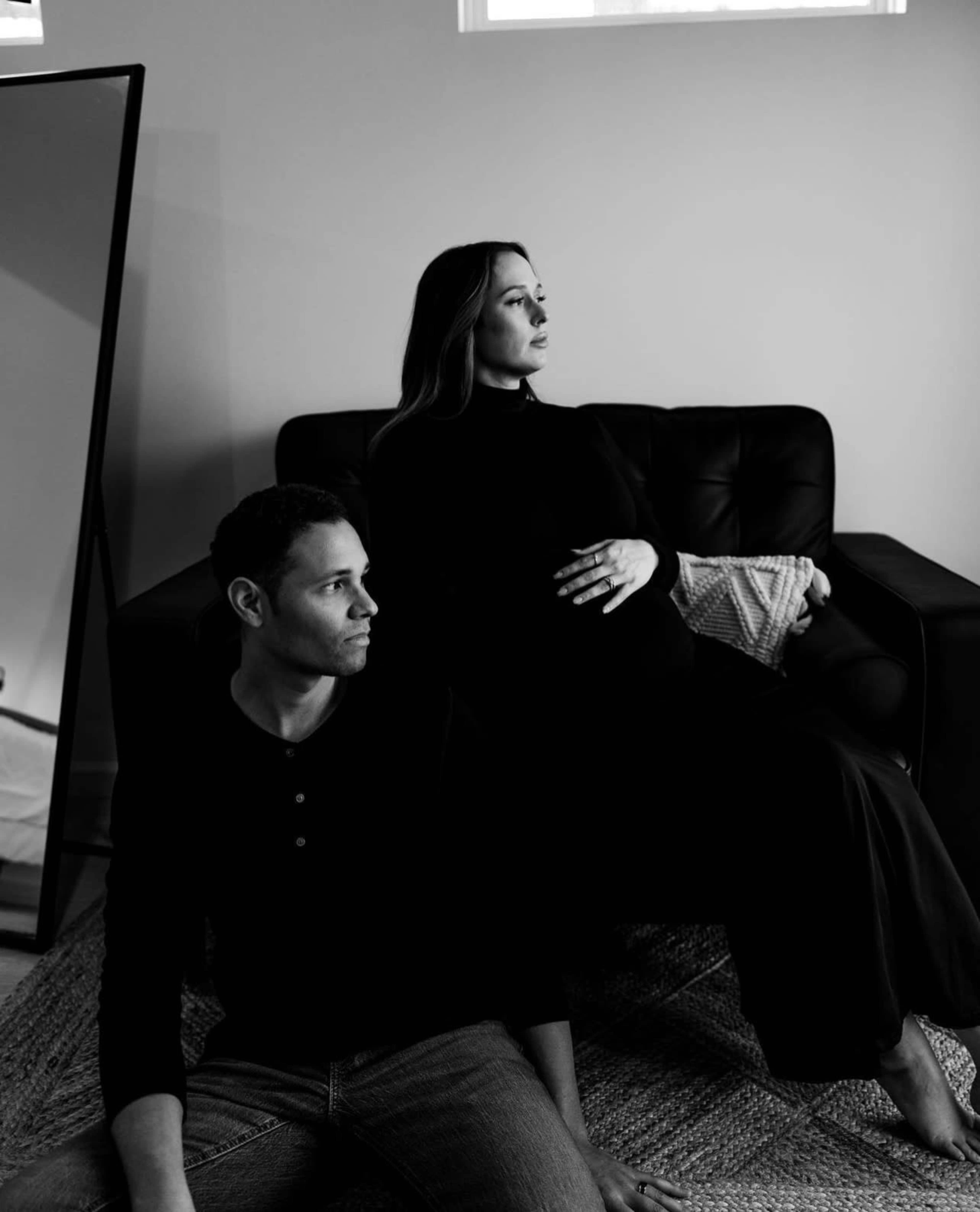 A pregnant woman in a black dress sits on a couch while a man in a black shirt gazes away, both positioned within a softly lit room.