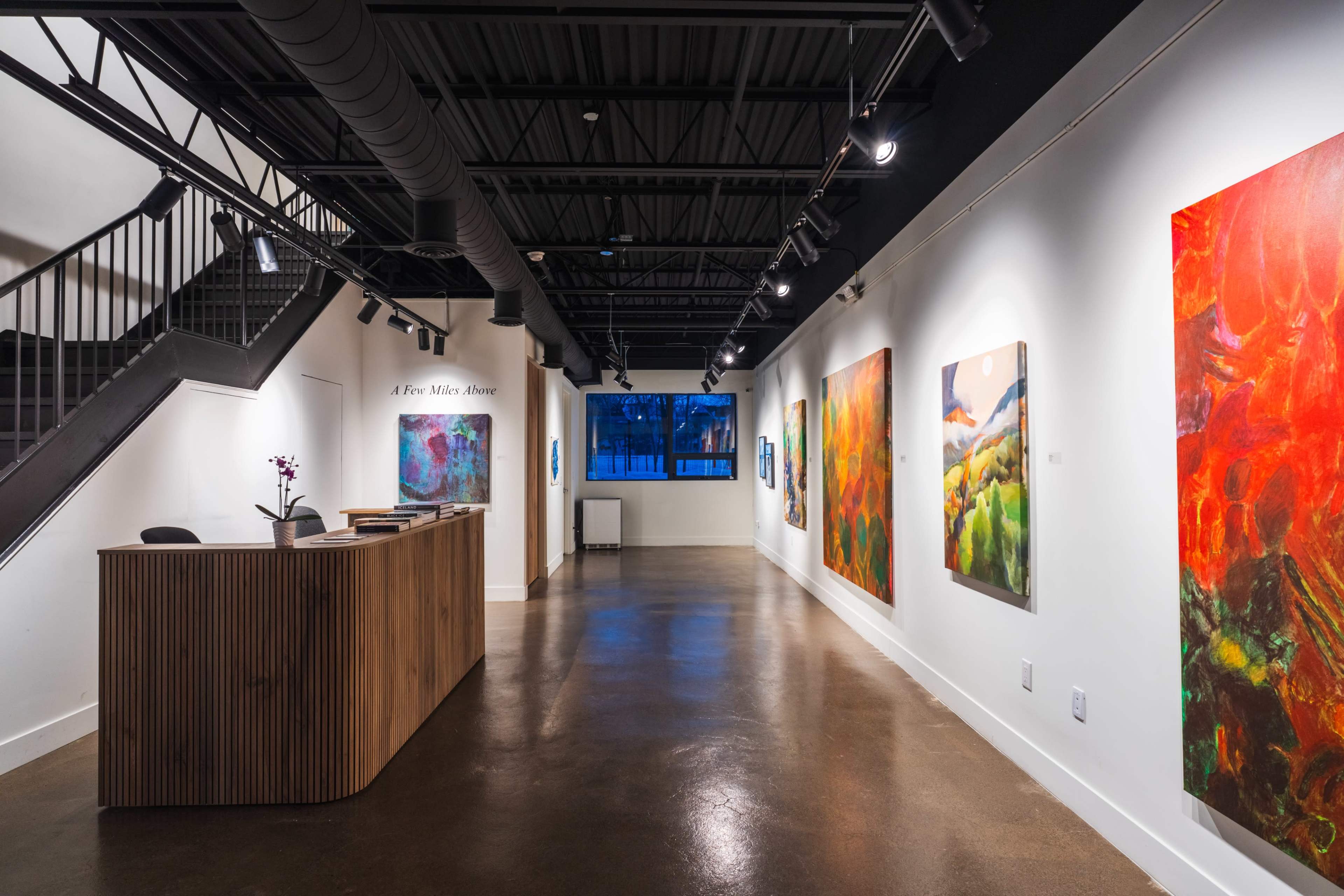 The image shows the interior of an art gallery with colorful abstract paintings on the walls, a reception desk on the left, and a staircase leading to an upper level.