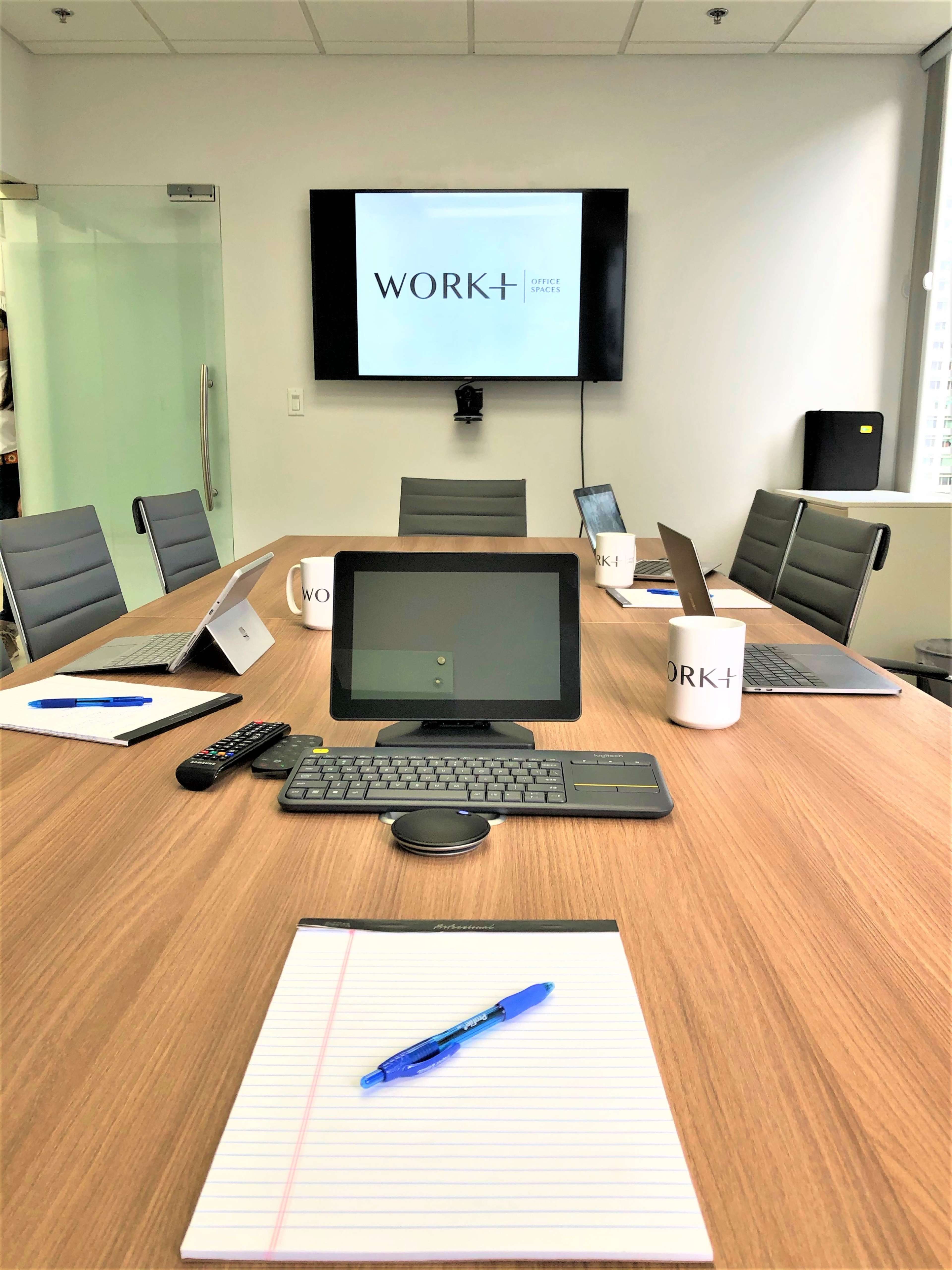 A conference room is set up with a wooden table, multiple laptops, a digital display showing the word "WORK+", and stationary items including notepads and pens.