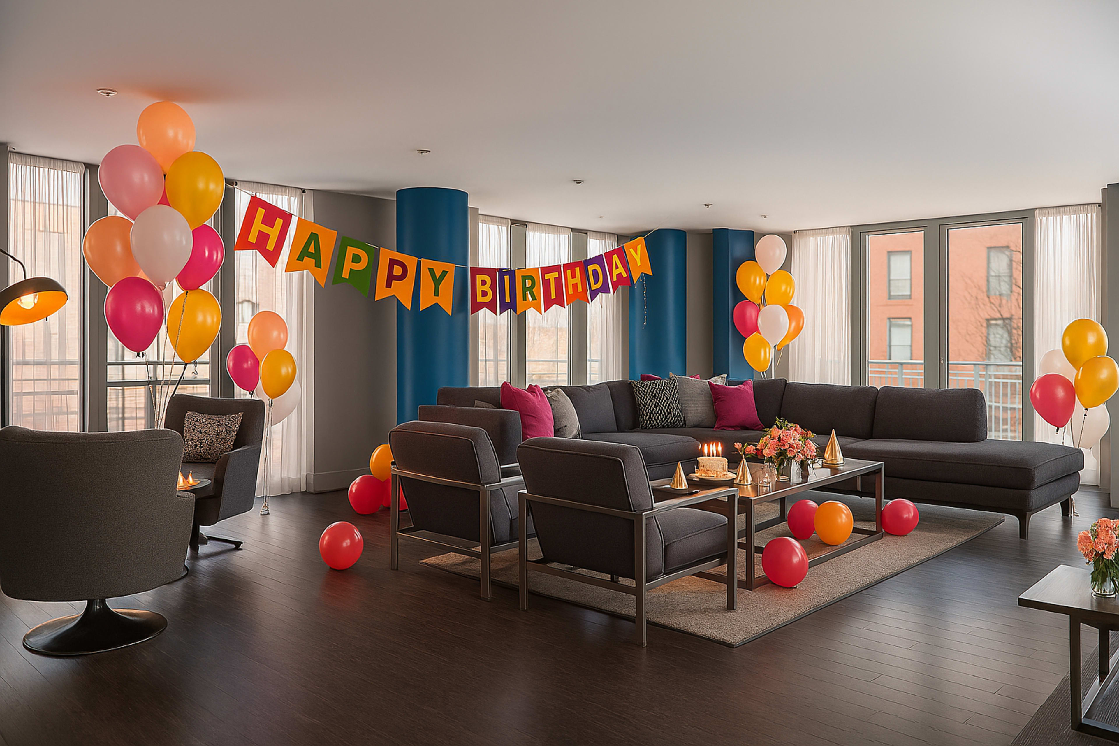 A living room is decorated with colorful balloons and a "HAPPY BIRTHDAY" banner, featuring a seating area with a dark gray sofa and armchairs.