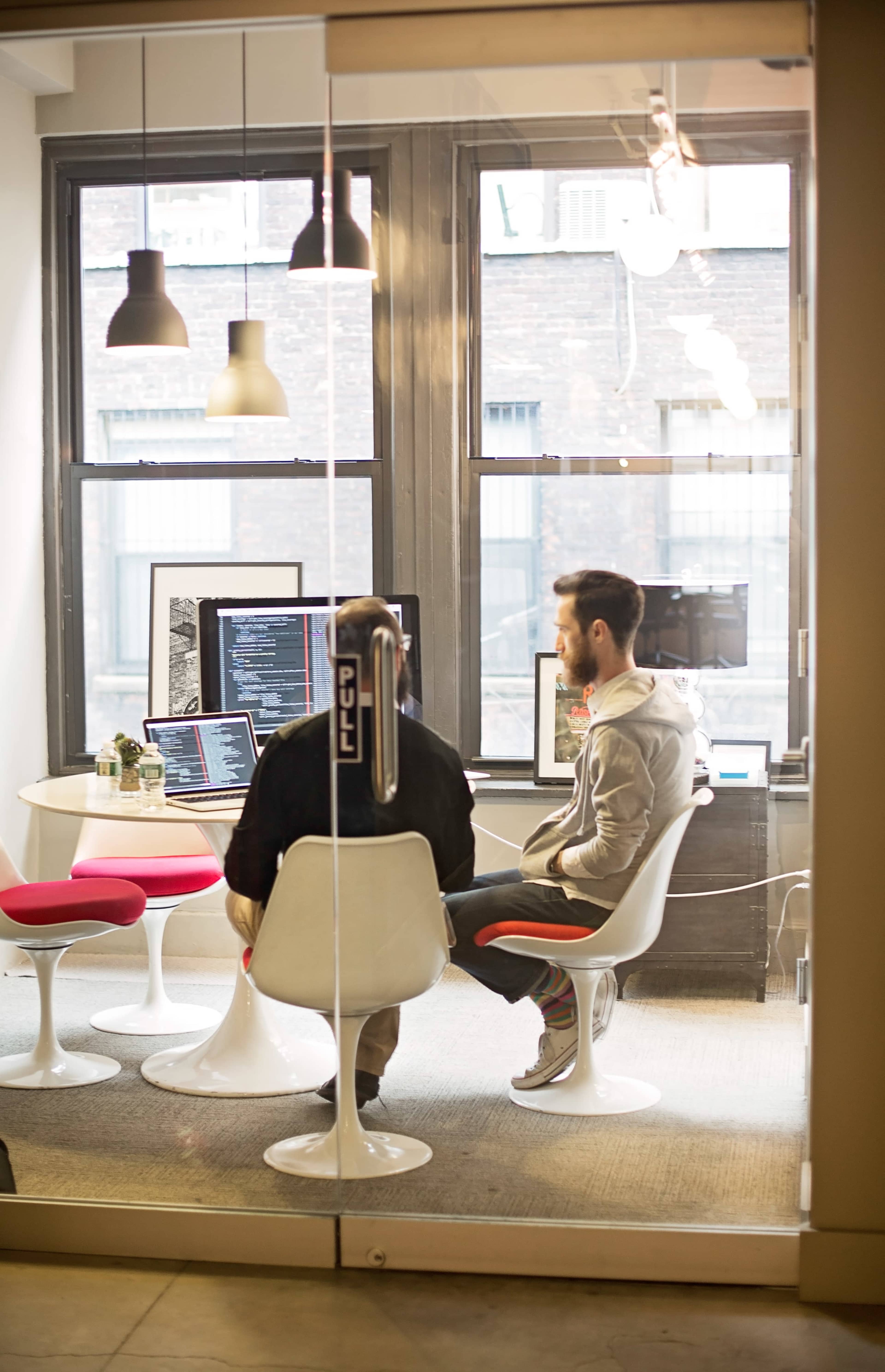 Two people sit in a glass-walled meeting room, discussing information displayed on their computer screens.