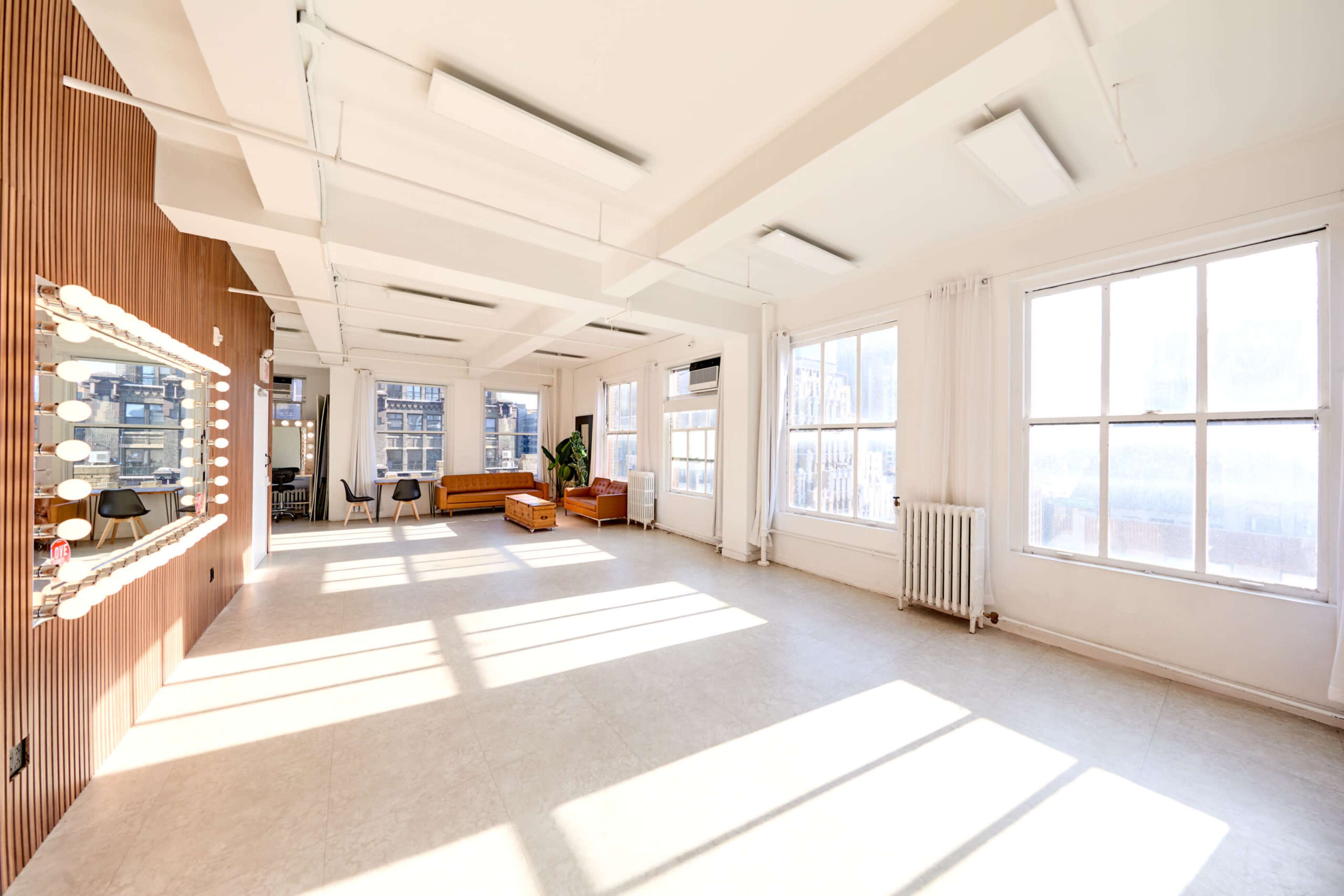 The image shows a spacious, well-lit room with large windows, a wooden accent wall featuring vanity lights, and minimalist furniture including a couch and decorative plants.