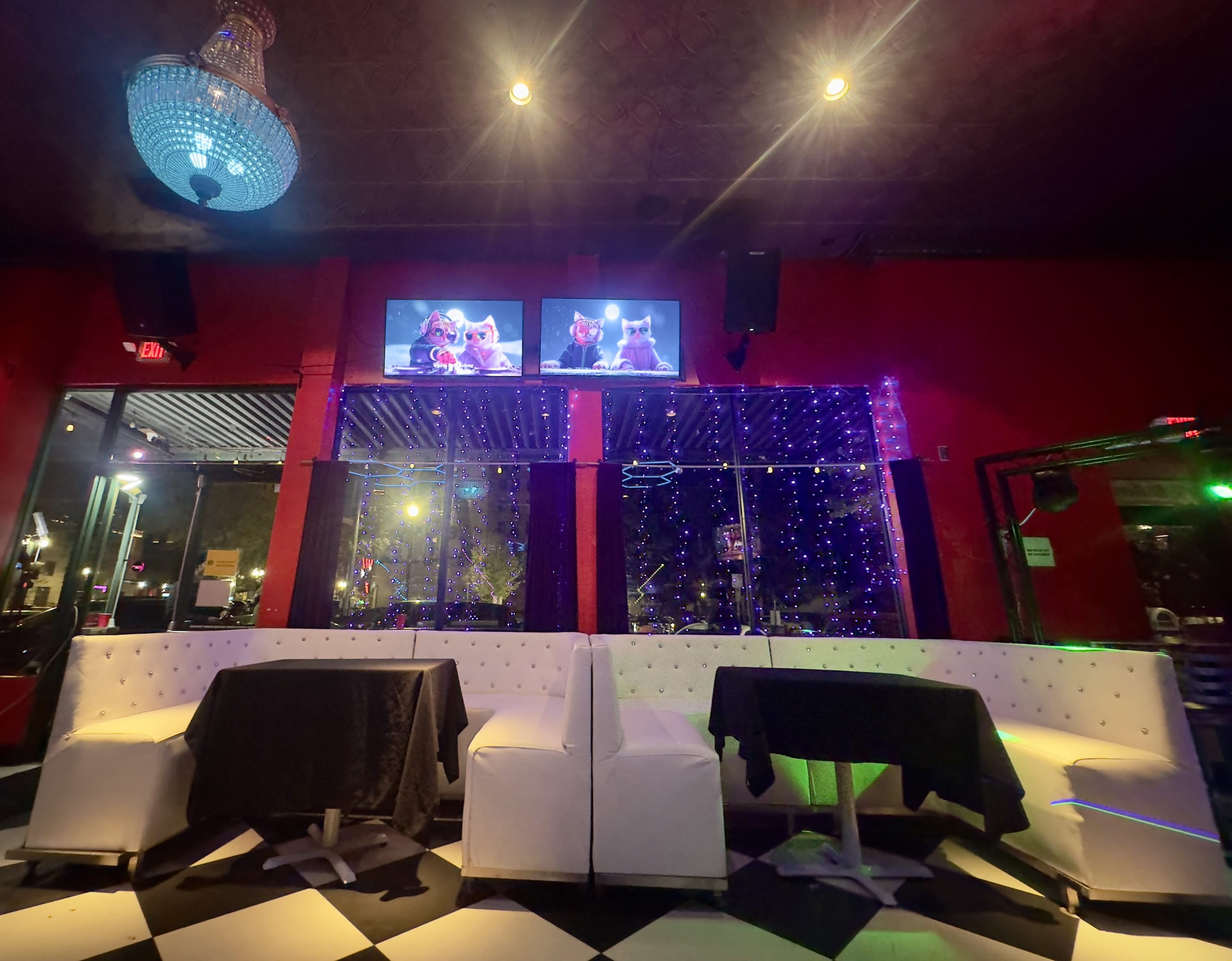 The image shows a sleek lounge area with white seating, black table linens, and two screens displaying images, surrounded by colorful lights and a chandelier.