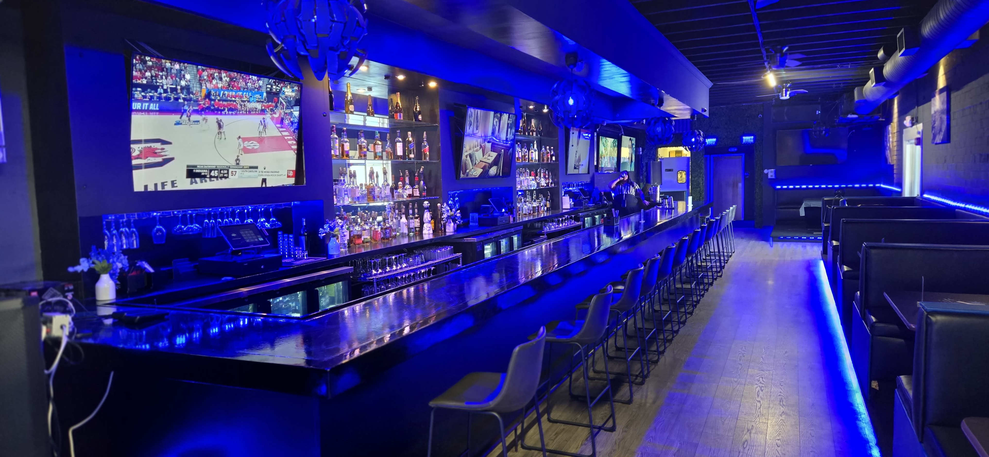 A dimly lit bar features a long mirror-topped counter, illuminated by blue lights, with shelves of liquor and a large screen displaying a basketball game in the background.