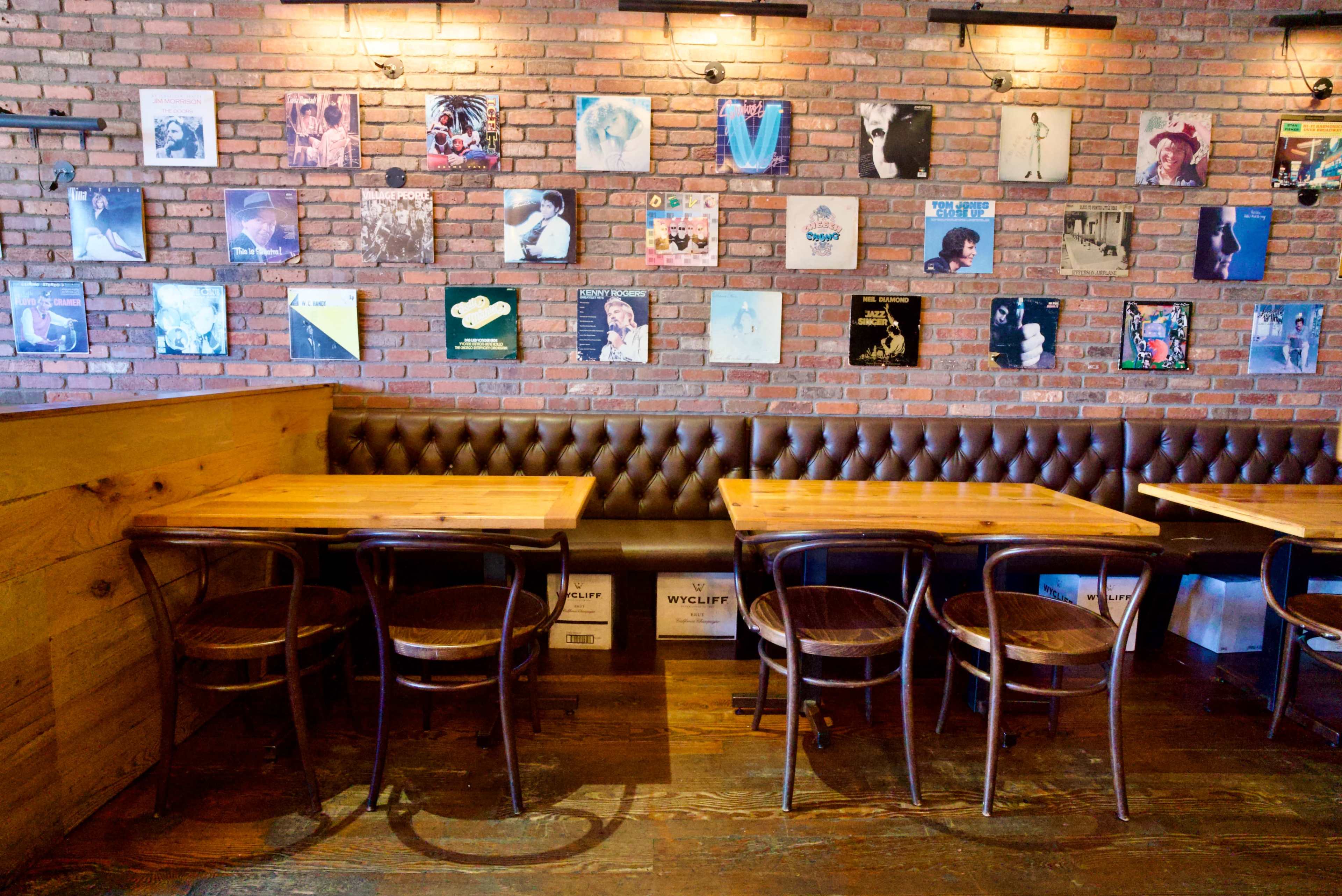 The image shows a cozy restaurant interior with wooden tables, leather-backed seating, and a brick wall adorned with various vinyl record covers.