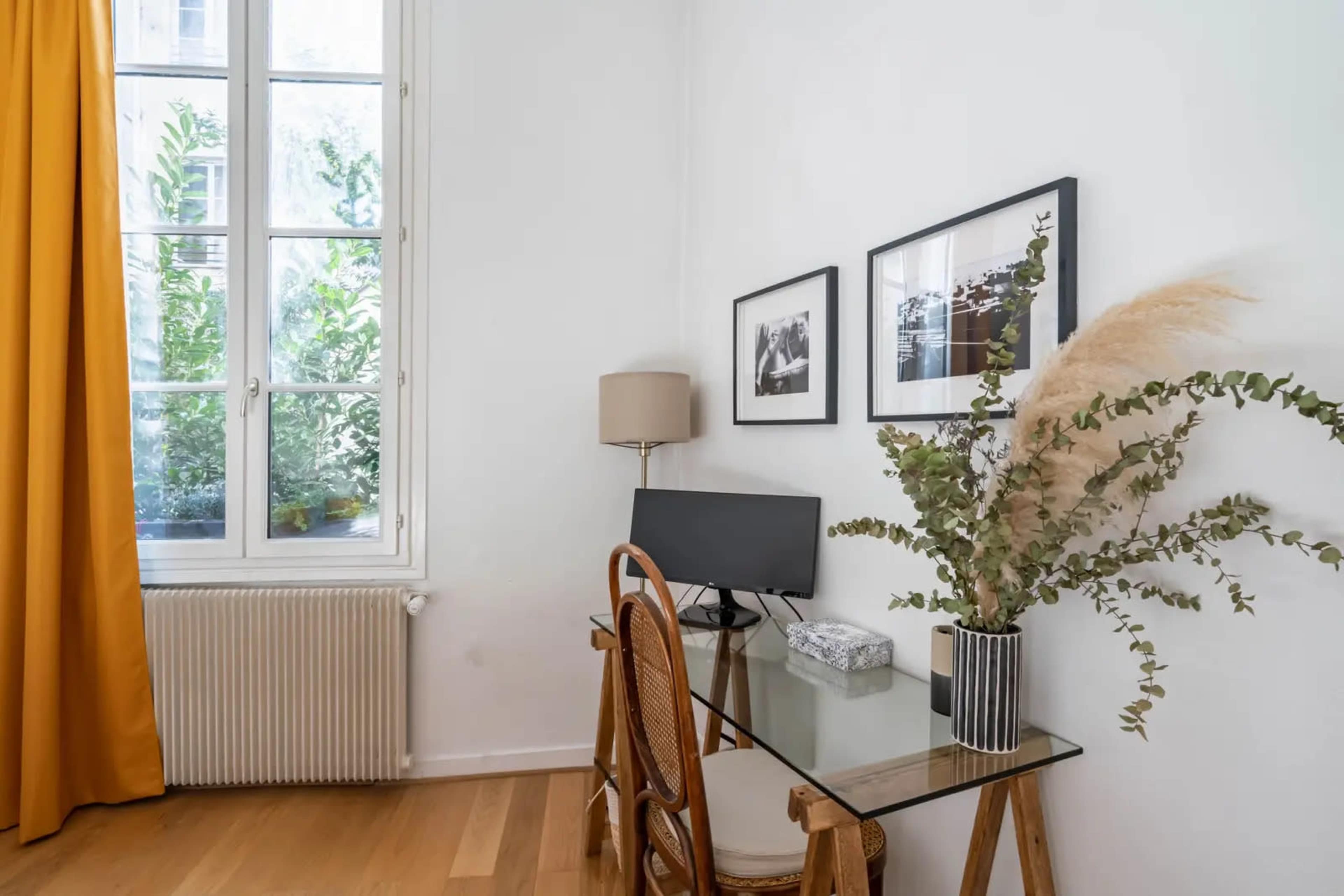 A small workspace features a glass desk with a computer, a lamp, and decorative plants, beside a window dressed in yellow curtains.