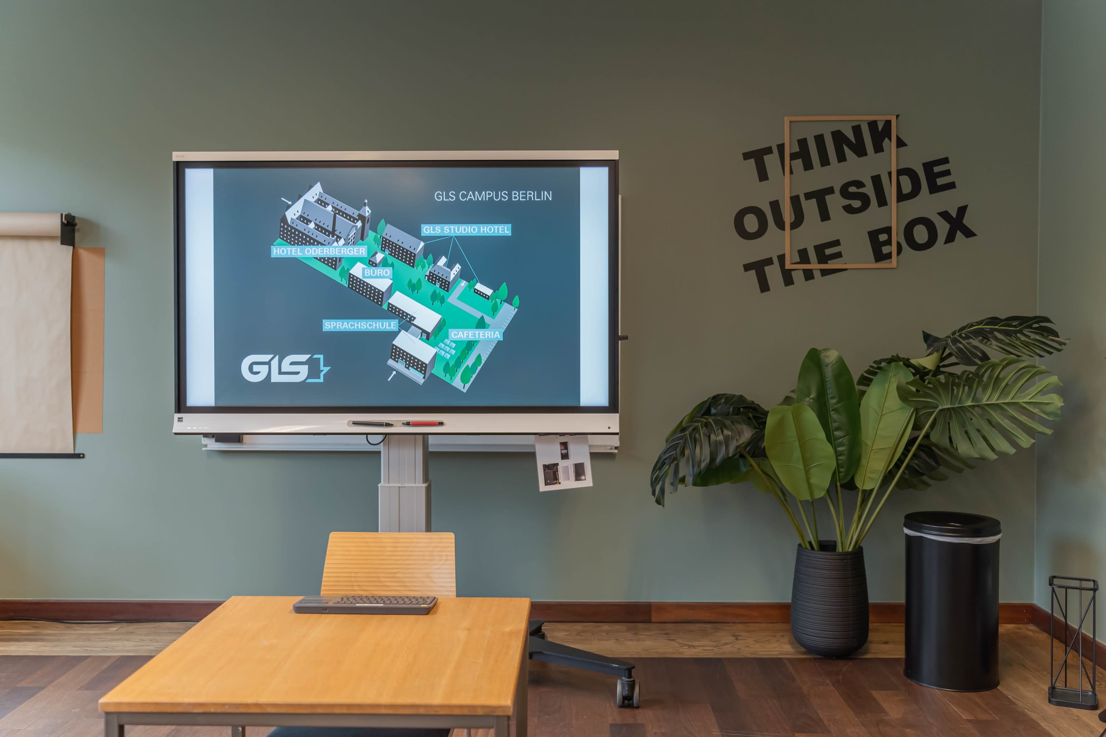 The image shows a modern meeting room featuring a digital display of the GIS Campus Berlin layout, a wooden table with a laptop, and a plant beside a wall with a motivational phrase.