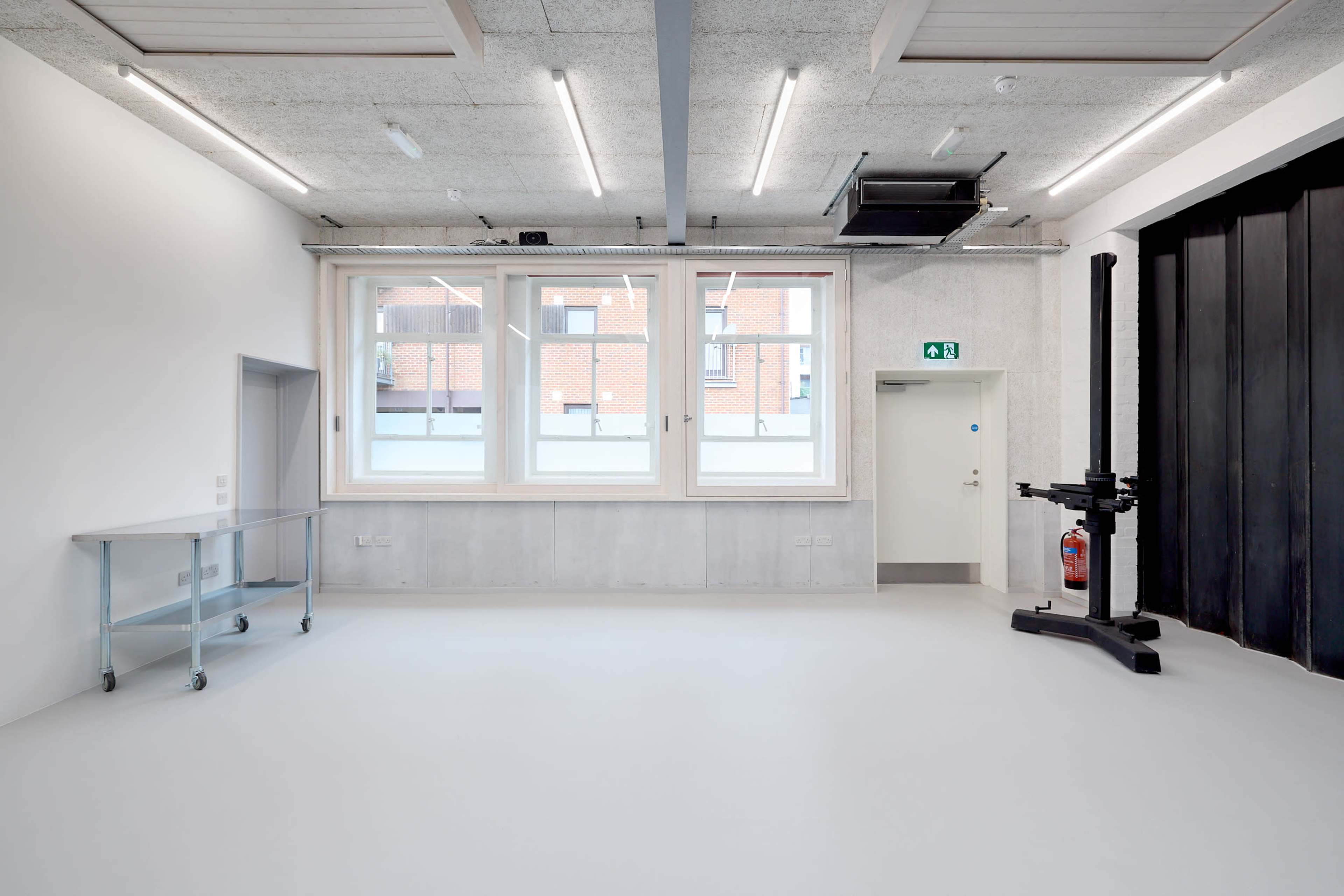 The image shows a spacious, minimally furnished room with large windows, a gray concrete wall, and a large black equipment stand in one corner.