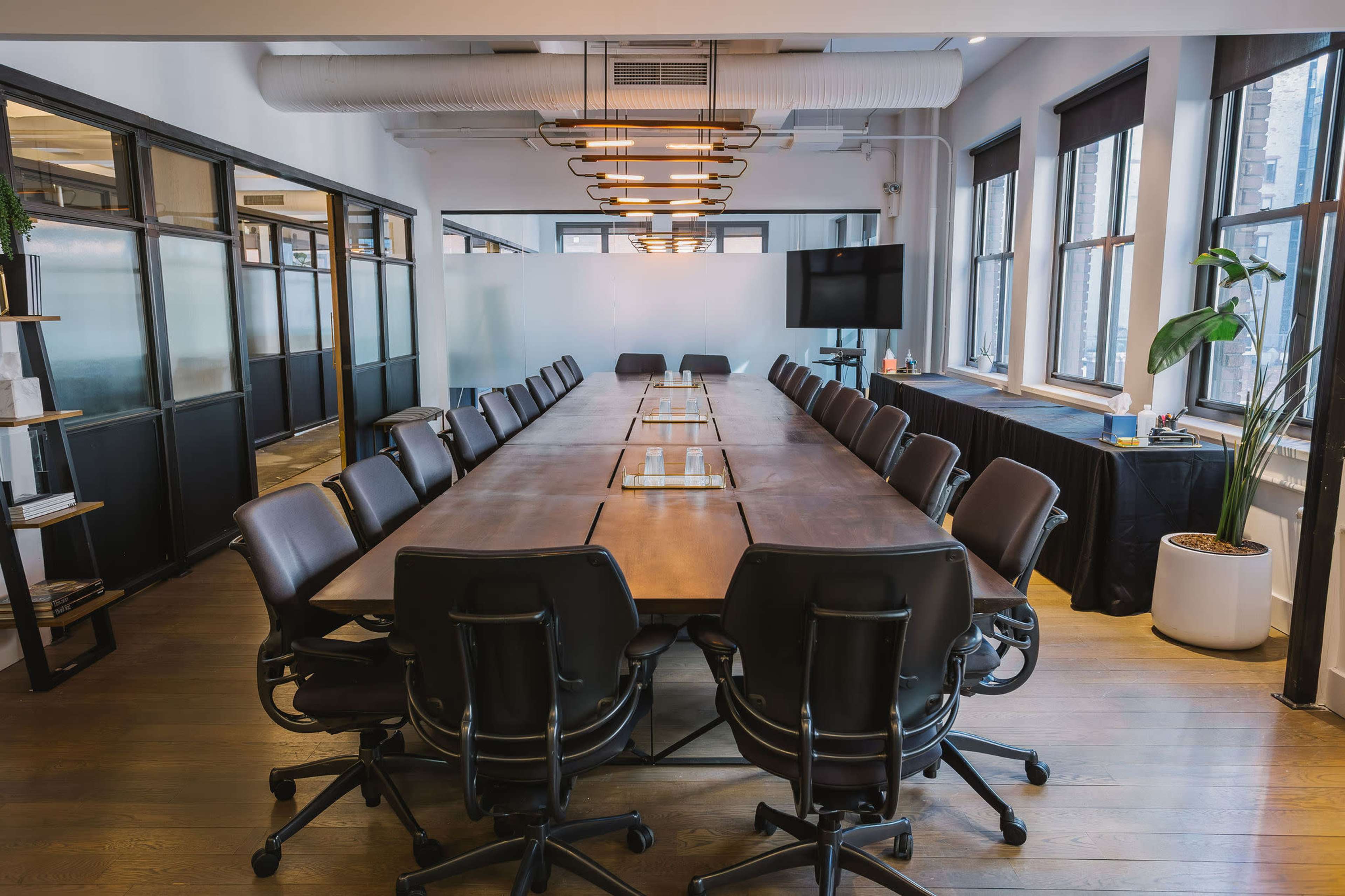 A large conference room features a long wooden table surrounded by ergonomic black chairs, with natural light coming through windows and modern lighting overhead.
