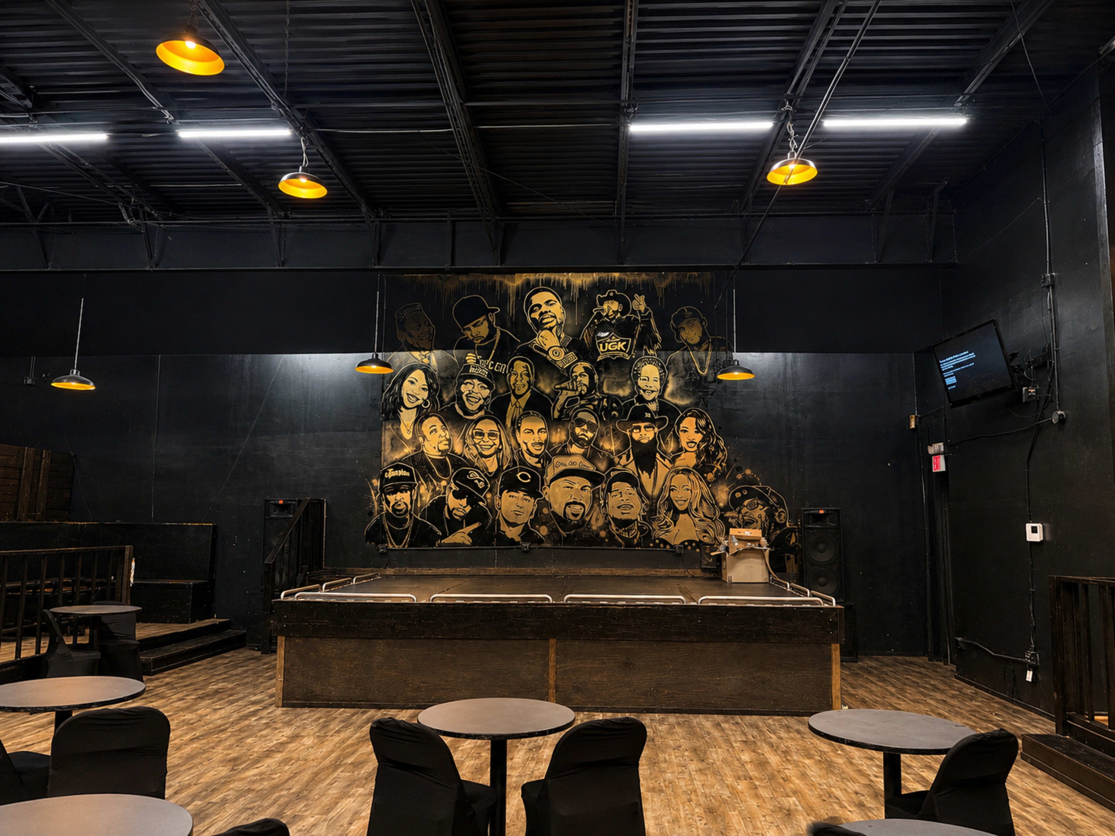 The image shows a stage in a venue with a large mural of various artists on the back wall, surrounded by tables and chairs facing the performance area.