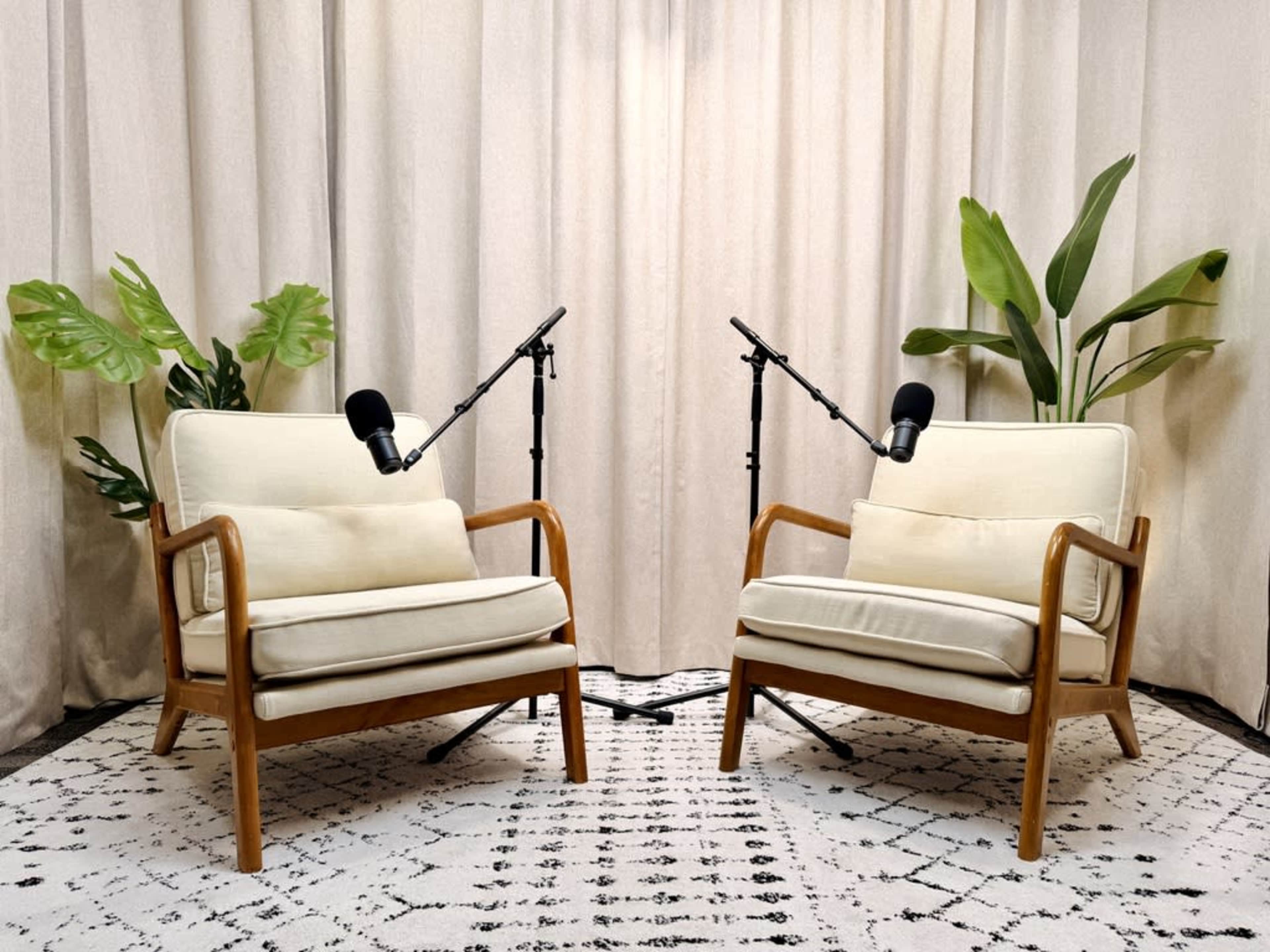The image shows two mid-century modern chairs facing each other with microphones on stands positioned between them, set against a light curtain and a patterned rug.