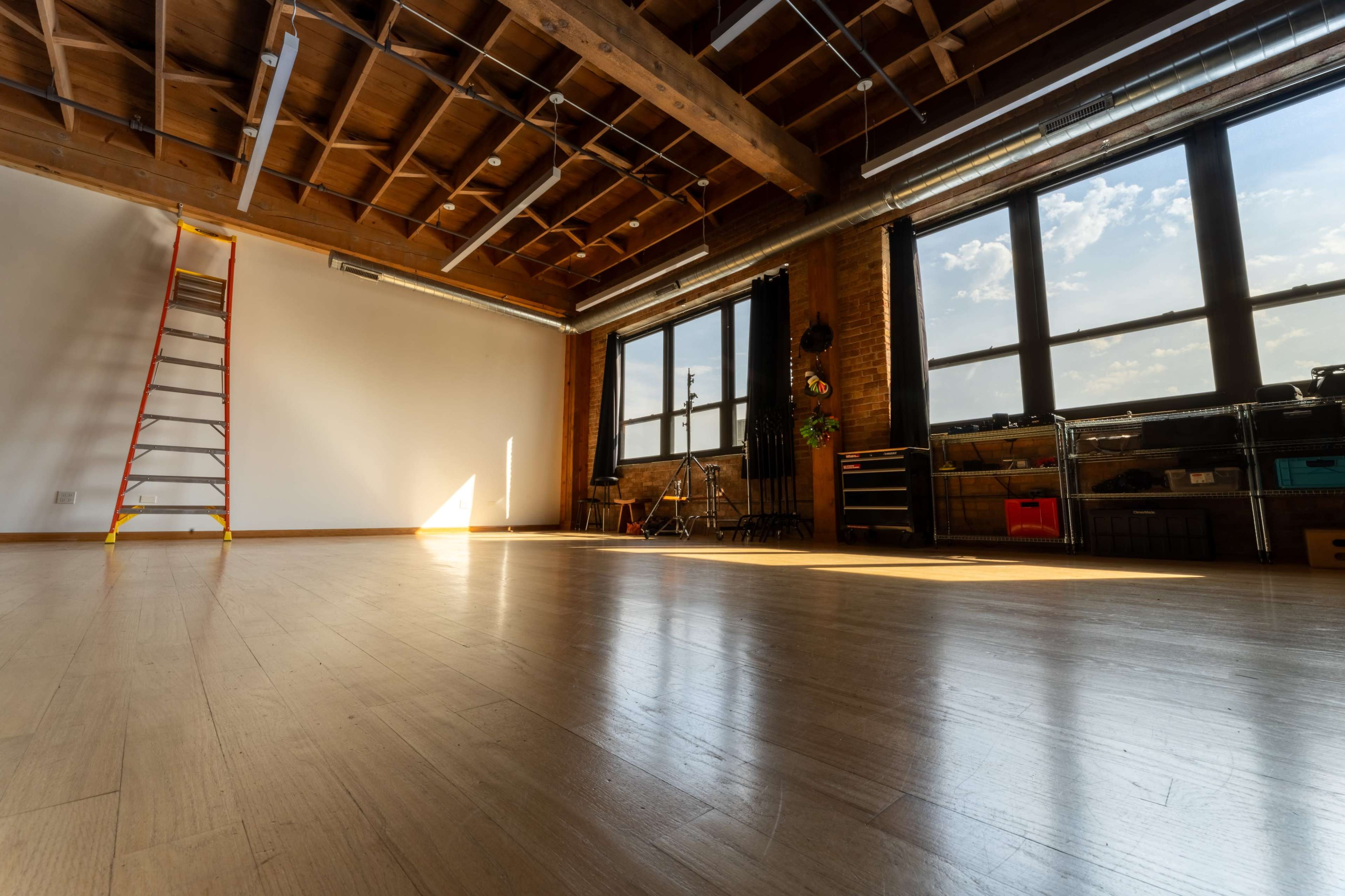 An empty room with wooden floors, large windows allowing daylight in, and a ladder leaning against one wall.