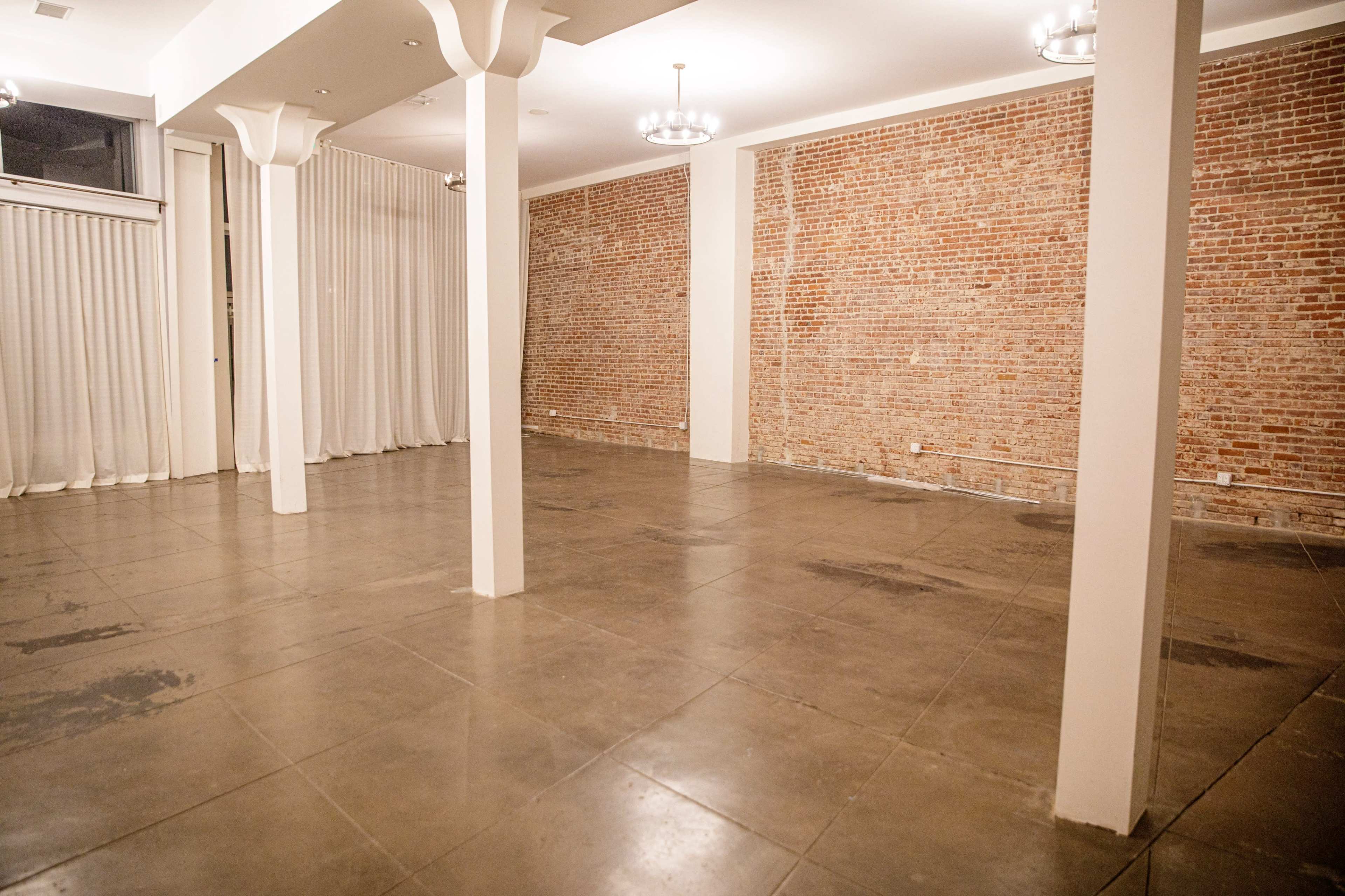 The image shows a spacious, empty room with exposed brick walls, concrete flooring, and several hanging light fixtures.