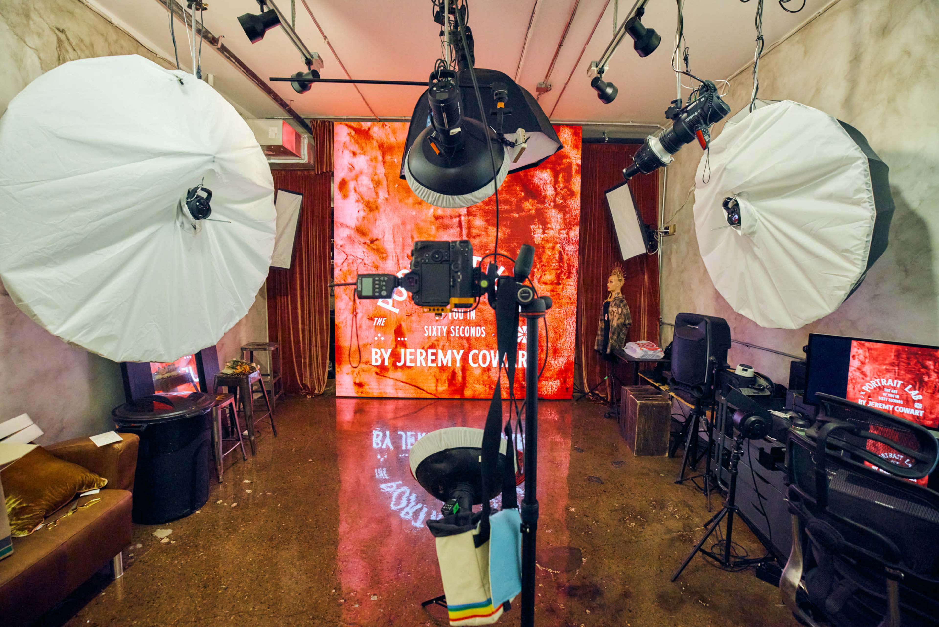 The image shows a photography studio set up with lights, softboxes, a camera on a tripod, and a backdrop displaying the text "THE SUNDAY SESSIONS BY JEREMY COVAR."