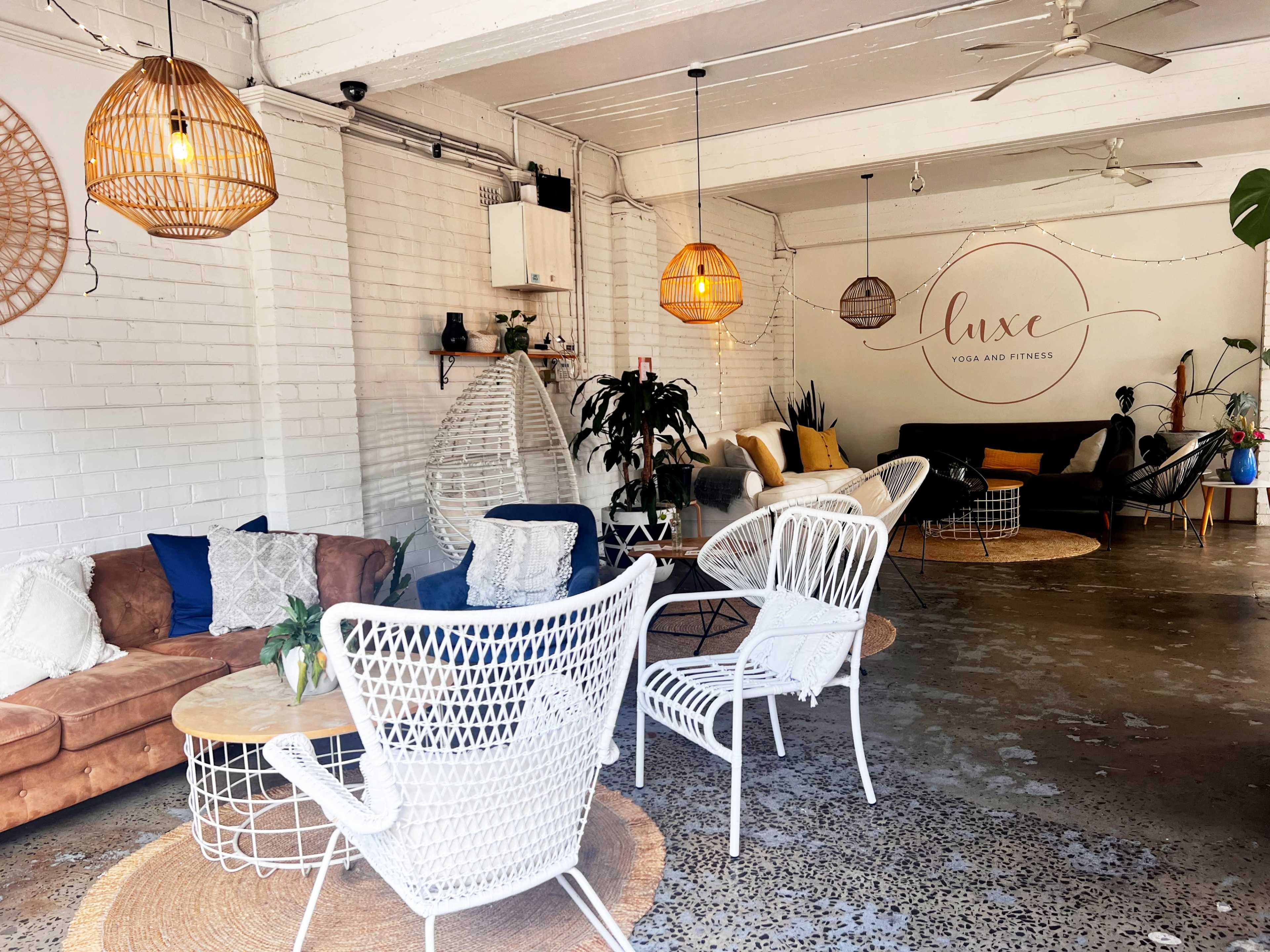 The image shows a cozy fitness and yoga studio with comfortable seating, decorative woven light fixtures, and plants in a bright, airy space.