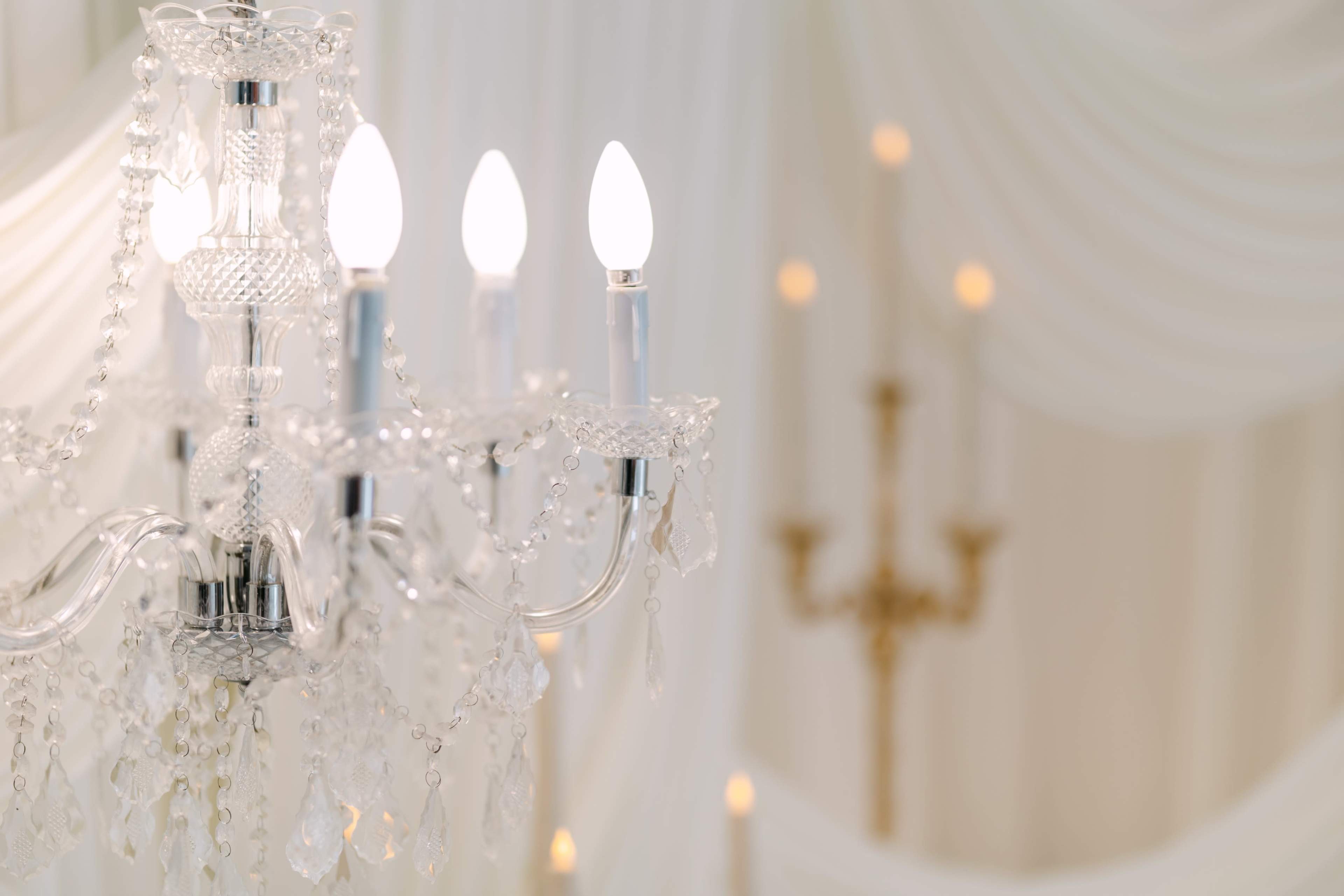 A crystal chandelier with three illuminated candle-like bulbs hangs in a softly lit room adorned with draped fabric and additional candlelight in the background.