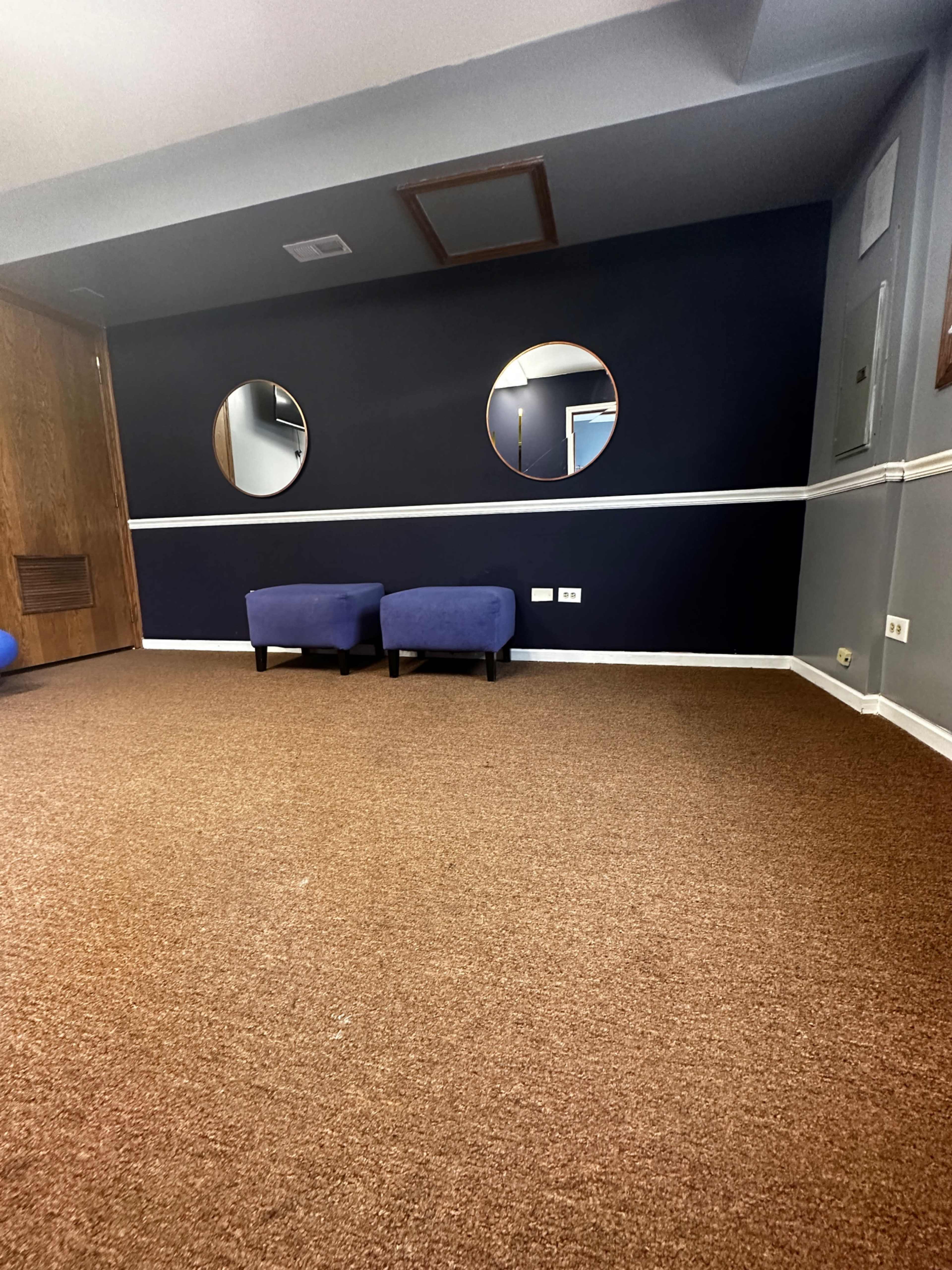 A room with brown carpet, two circular mirrors on a dark wall, and two blue stools near the center.