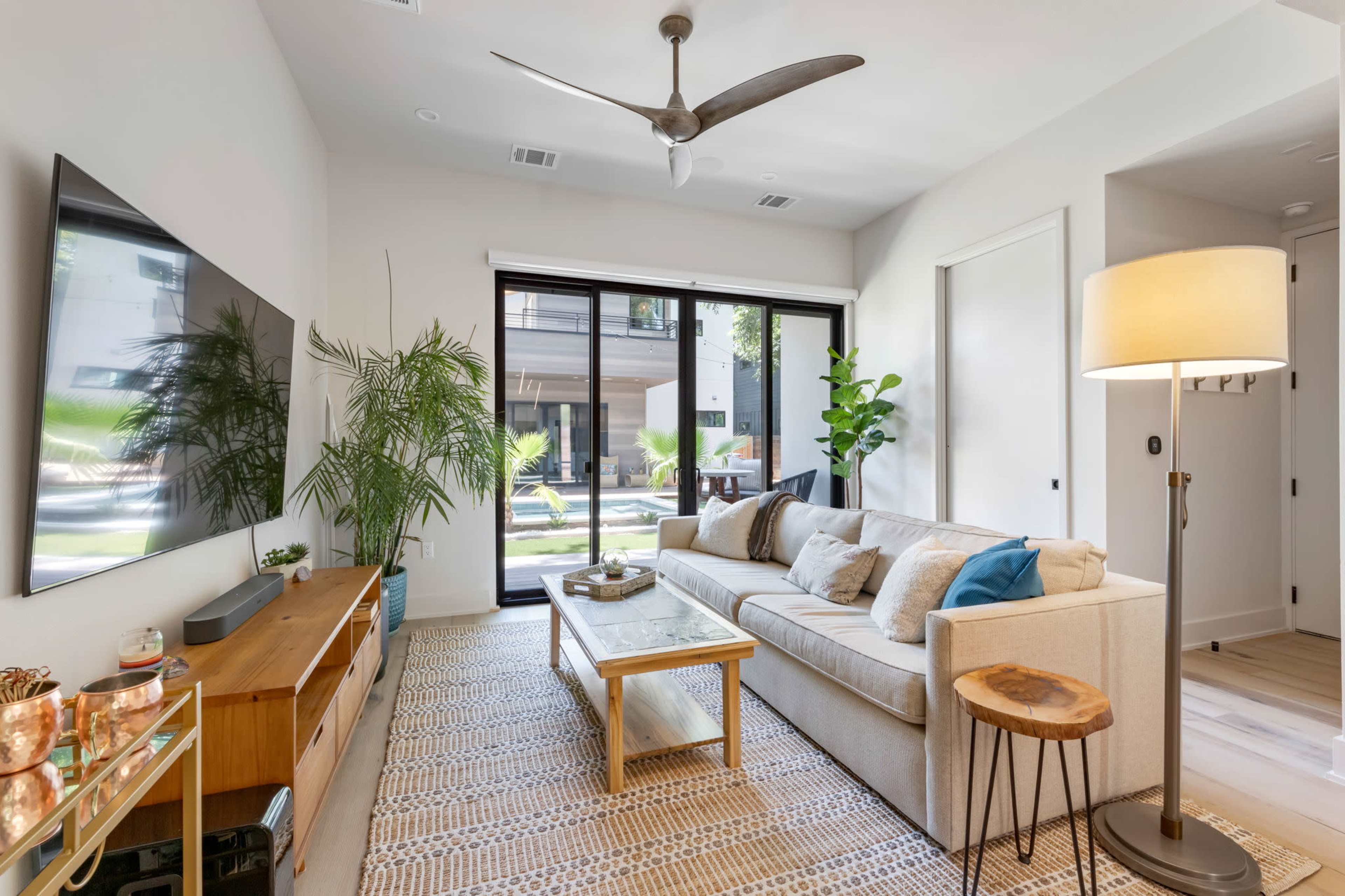 The image shows a modern living room with a light-colored sofa, a wooden coffee table, a ceiling fan, and large glass doors leading to an outdoor area.