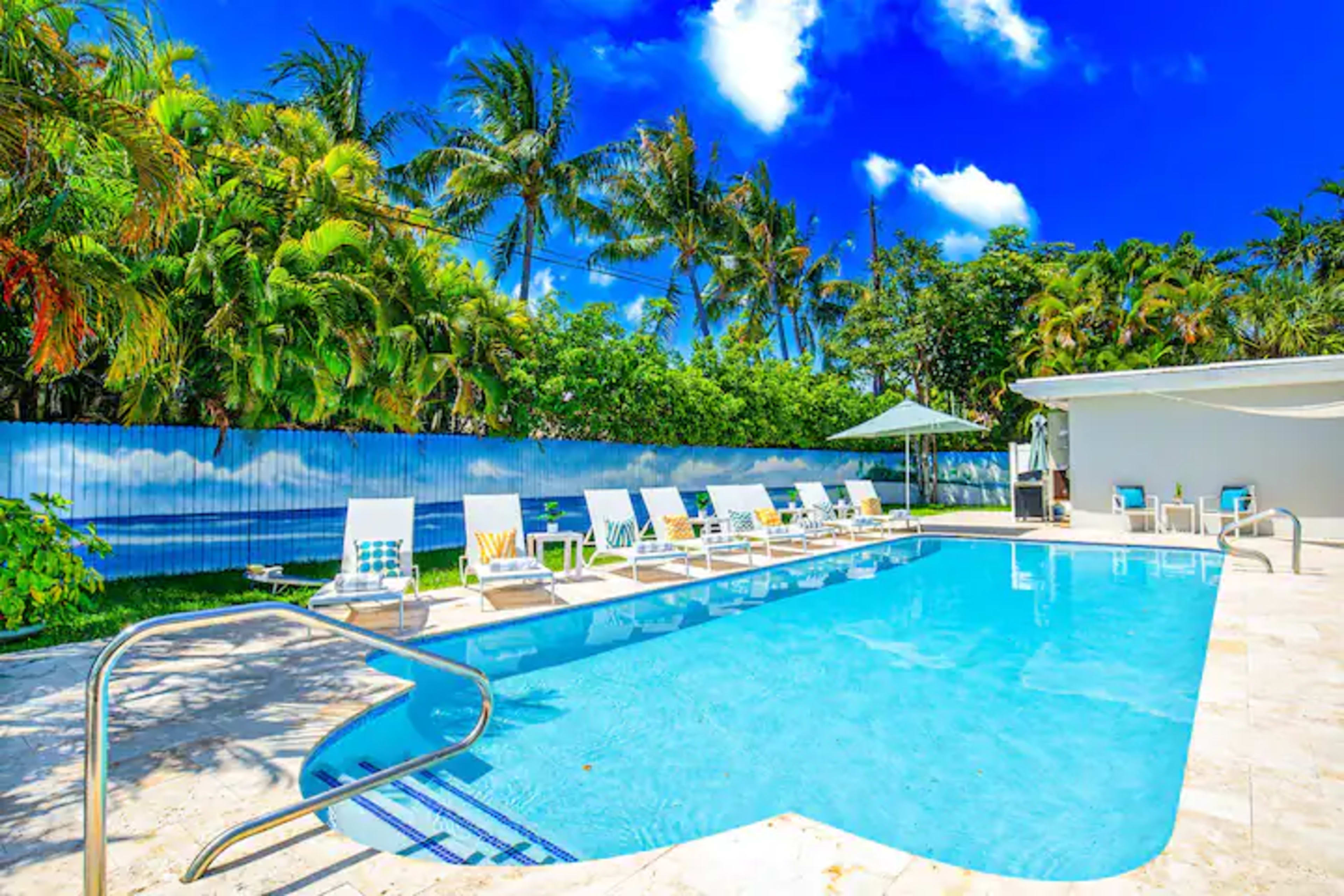 A swimming pool surrounded by lounge chairs and tropical greenery under a bright blue sky.