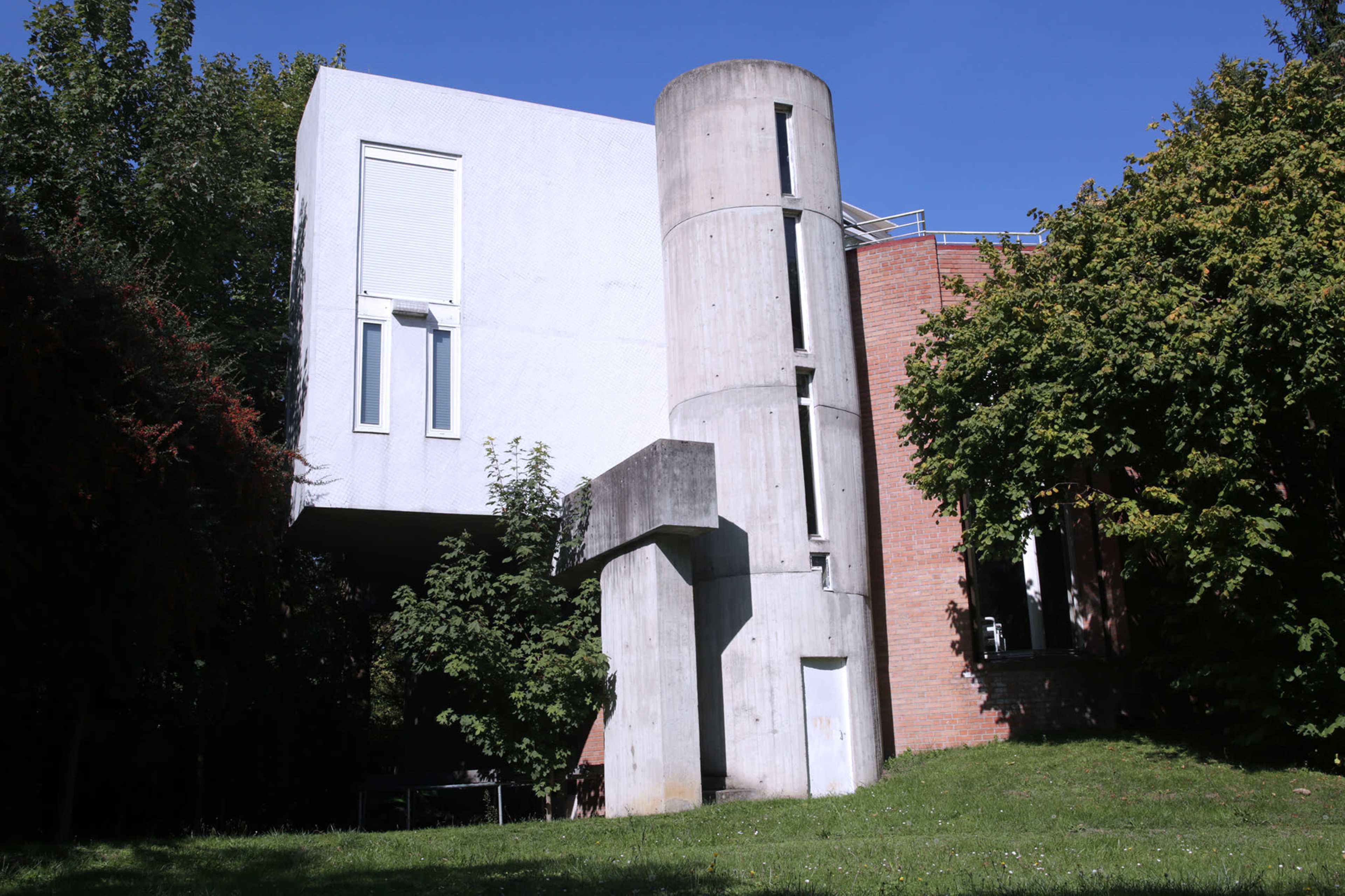 A modern two-story building featuring a mix of concrete and brick, with a cylindrical structure attached to one side and surrounded by greenery.