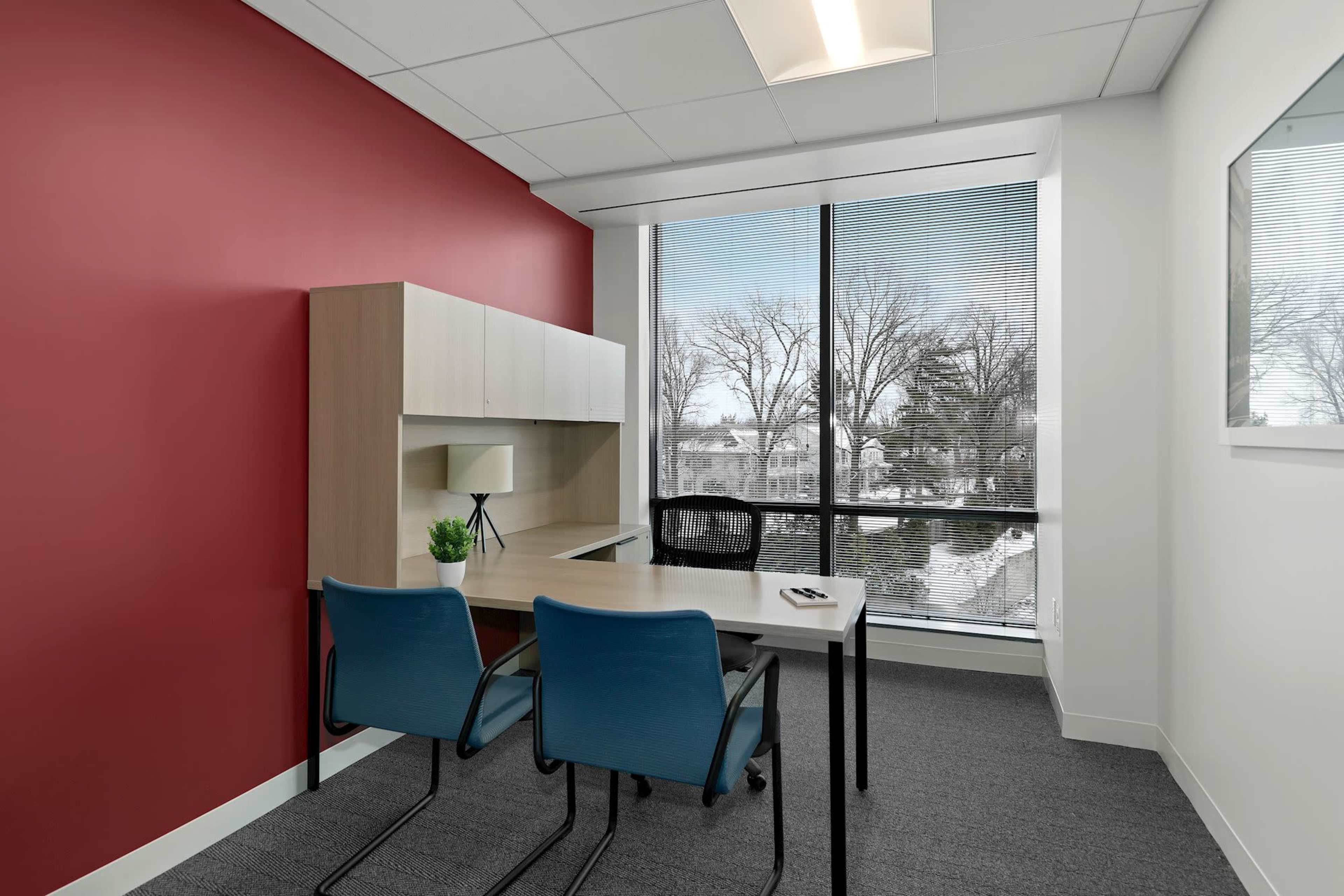 A bright office space featuring a desk, two blue chairs, and a window overlooking a snowy landscape with a red accent wall.