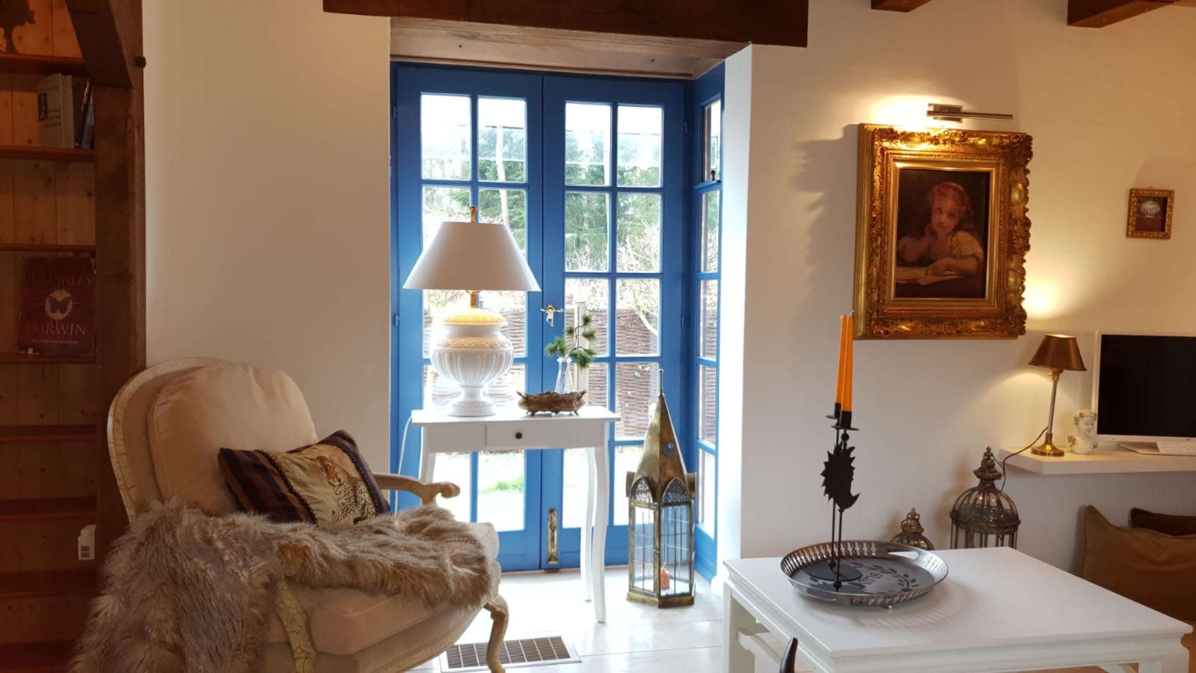 The image shows a cozy interior scene featuring a comfortable armchair, a small white table with a lamp, and blue French doors that open to a garden.