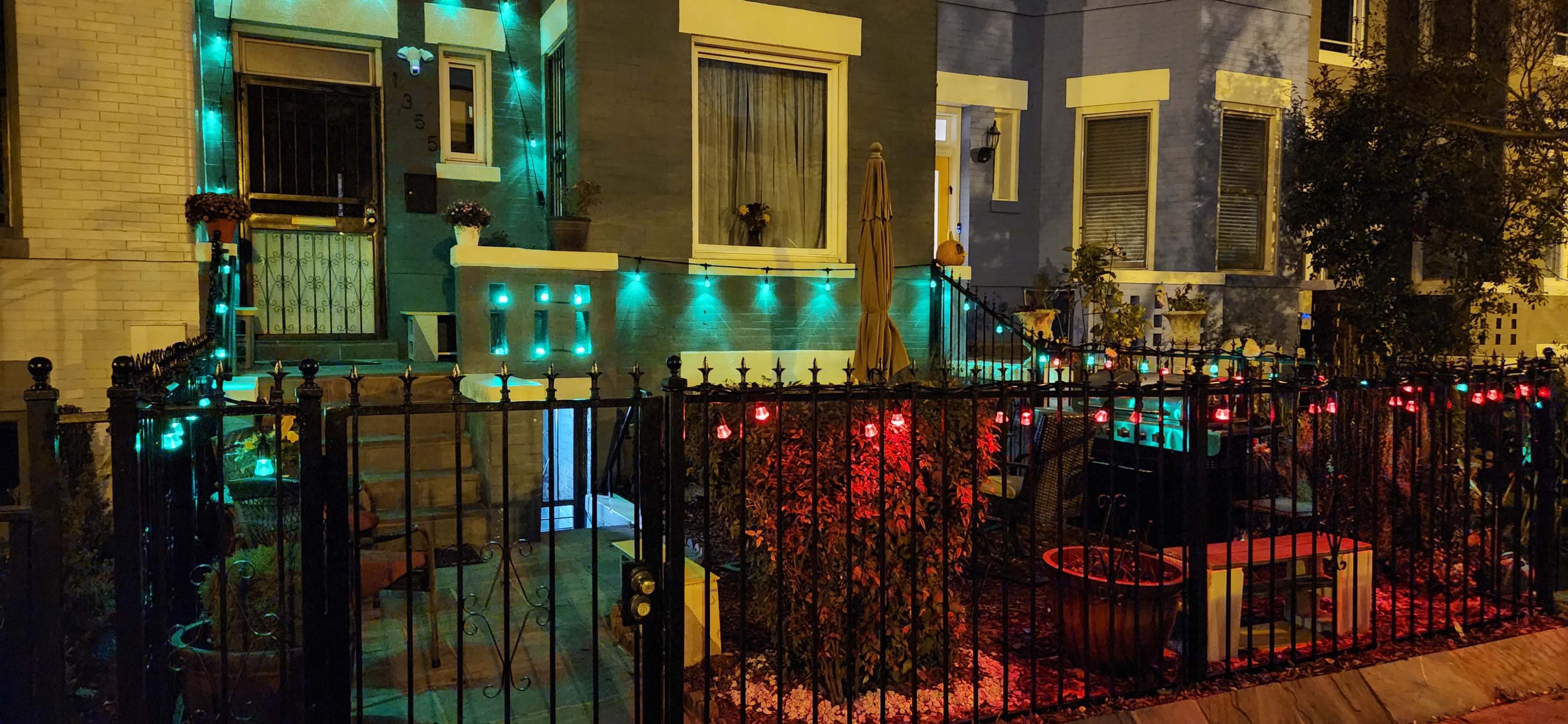 The scene shows a house adorned with green and red lights along the fence and entryway, with some potted plants and outdoor furniture in the yard.