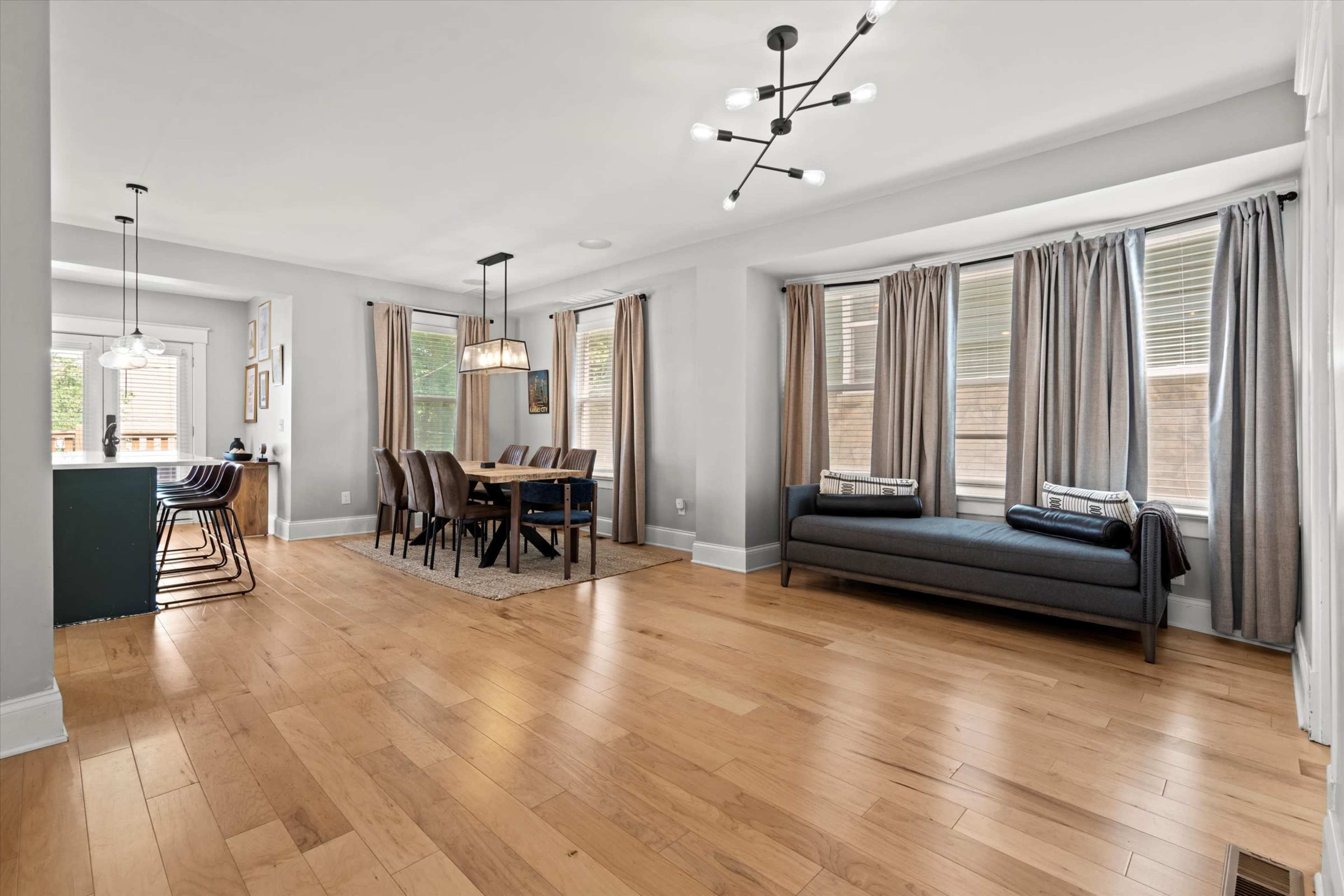 The image shows a modern living and dining area with hardwood floors, large windows covered by curtains, a dining table set for six, and a cozy seating area.