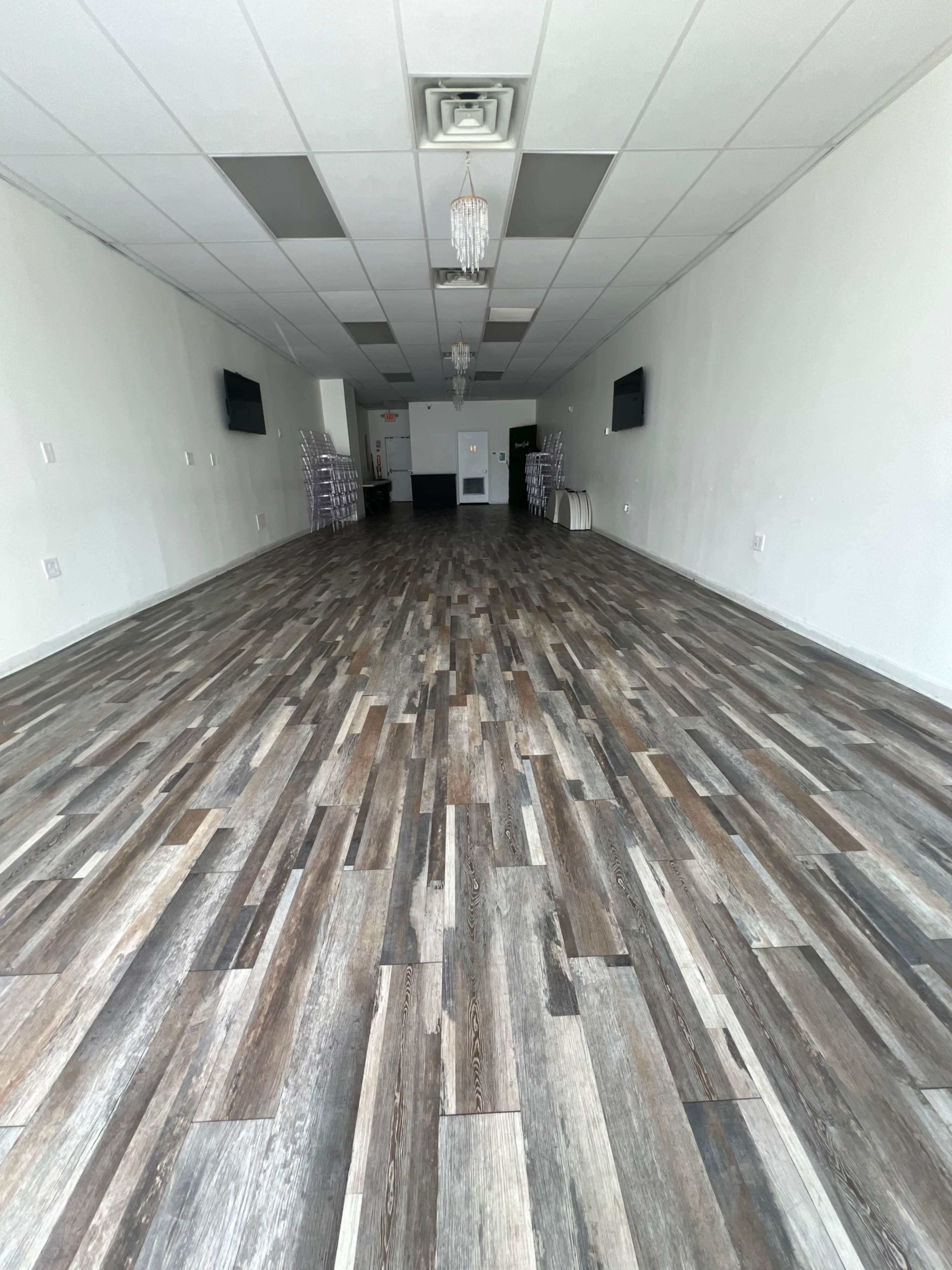 The image shows a spacious, empty room with wooden-style flooring, two flat-screen TVs mounted on the walls, and a few stackable chairs lined up against one side.