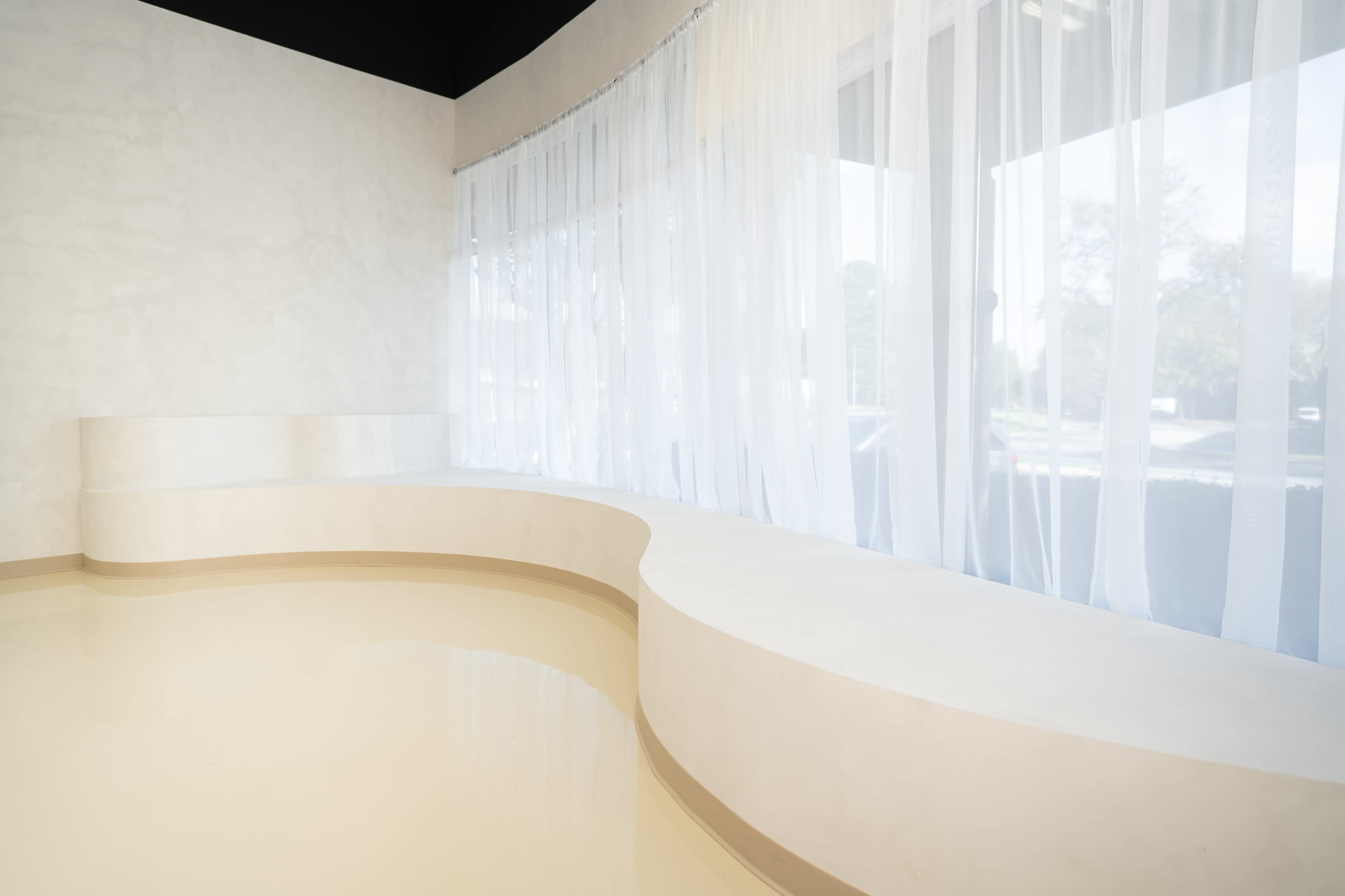 The image shows a curved white bench beside large windows draped with sheer curtains in a minimalist room.