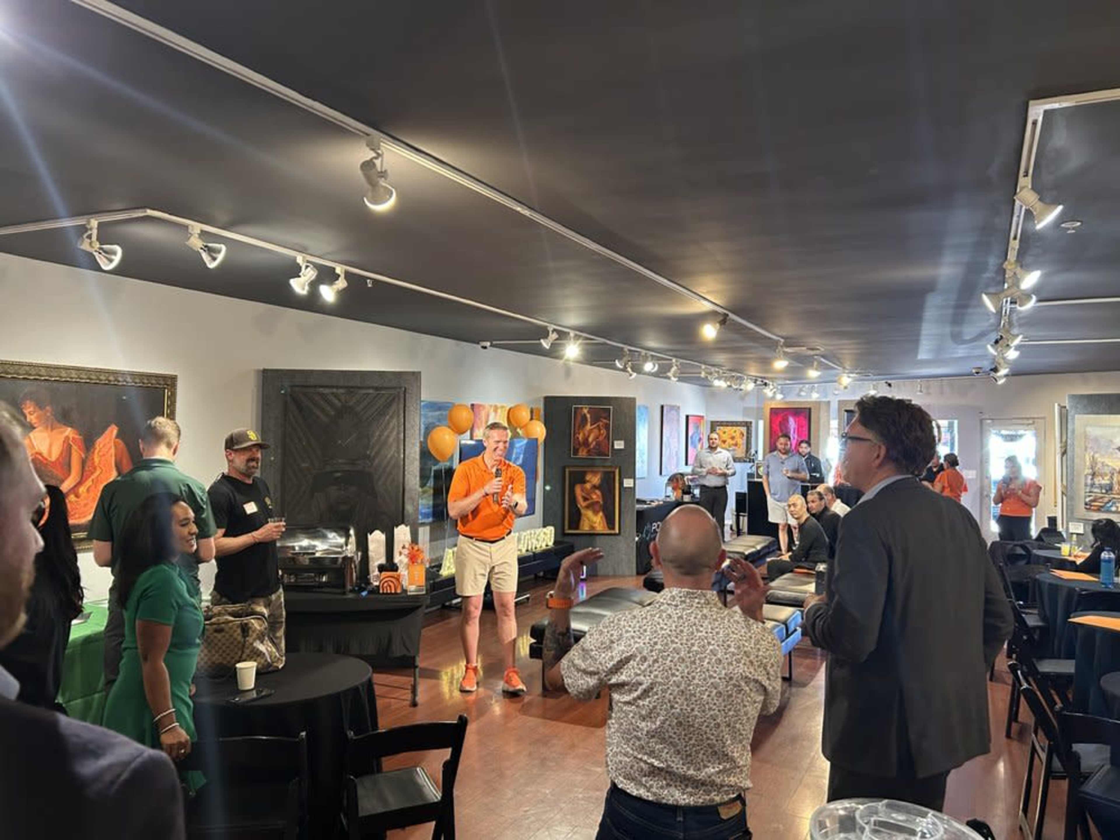 A group of people gathers in a gallery filled with art, where a speaker addresses the audience while others engage in conversation and enjoy refreshments.