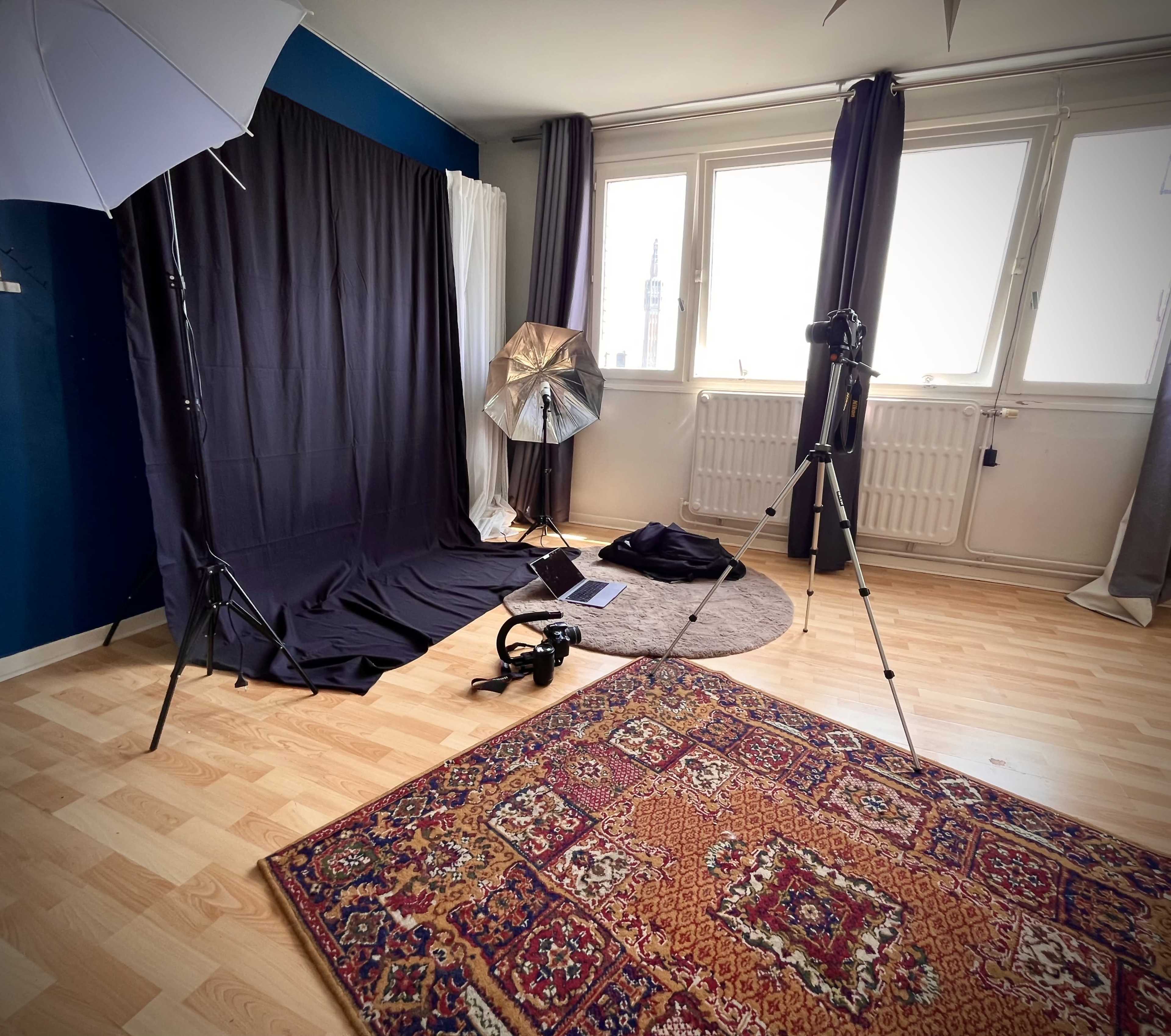 The image shows a photography studio setup featuring a black backdrop, lighting equipment, and a camera on tripods in a room with wooden flooring.