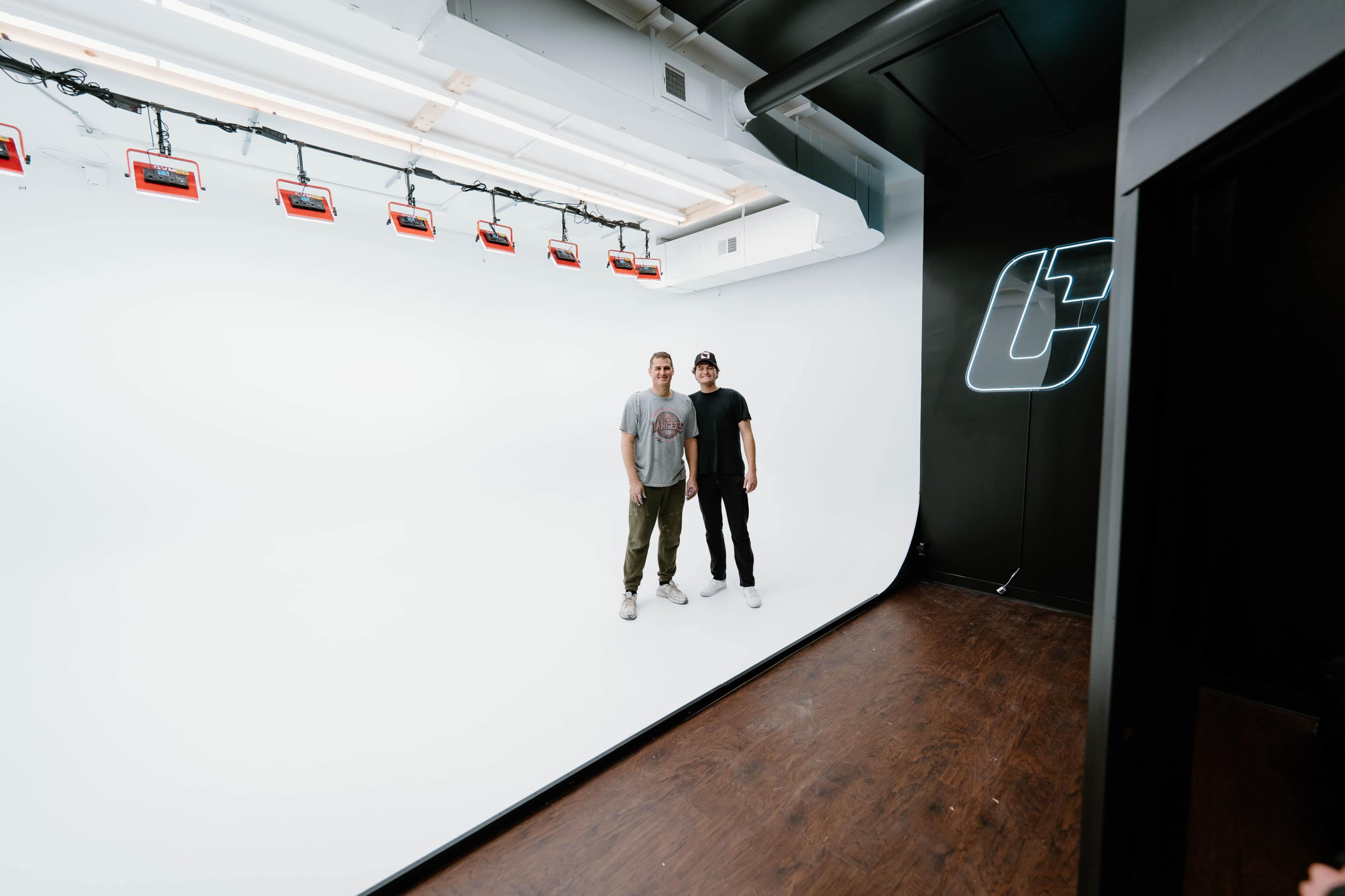 Two men stand on a wooden floor in a brightly lit white studio space with red lights hanging from the ceiling and a logo on the wall.