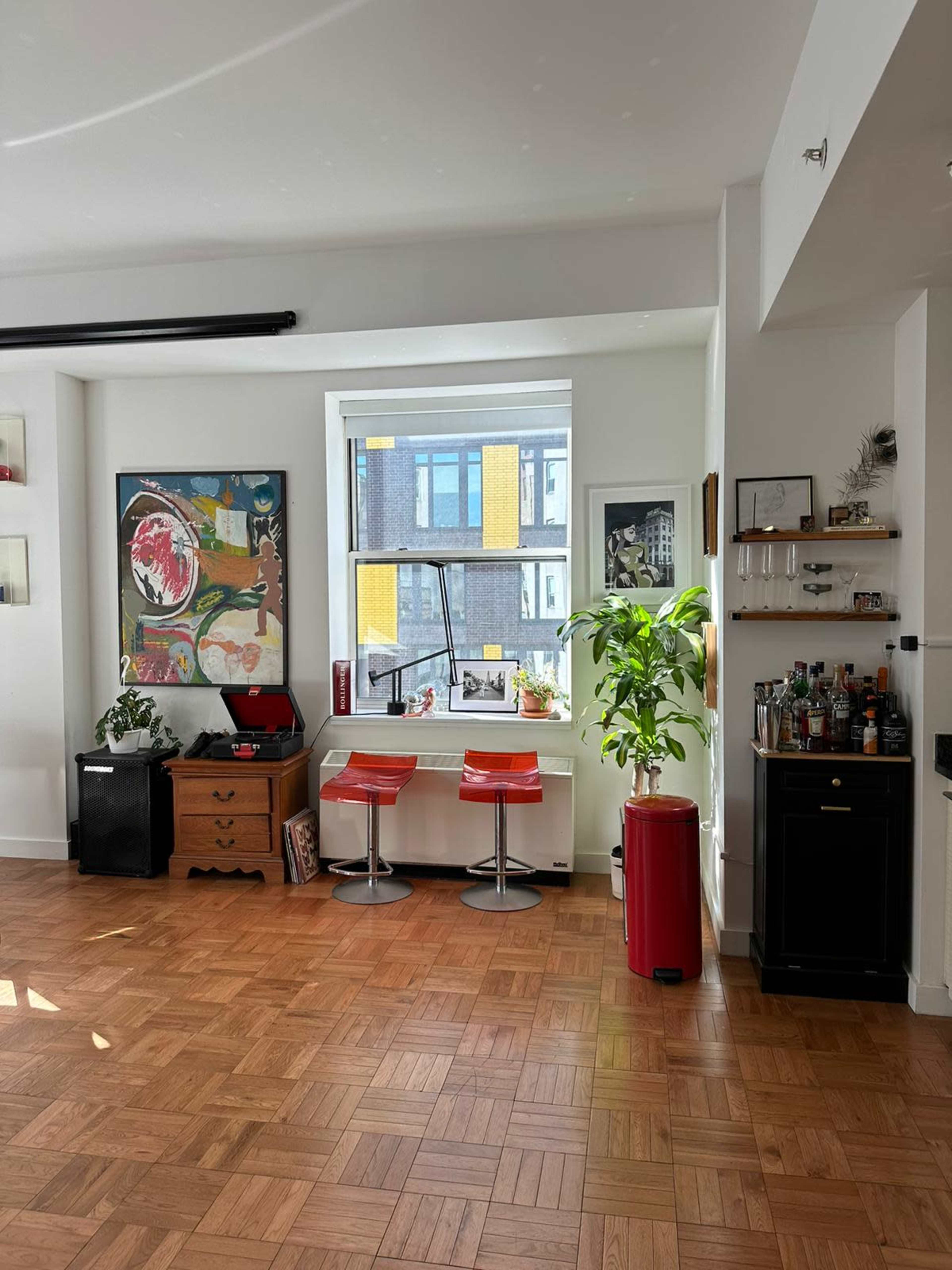 A bright room with two red bar stools positioned by a window, a wooden cabinet with art on the walls, and a bar cart in the corner.