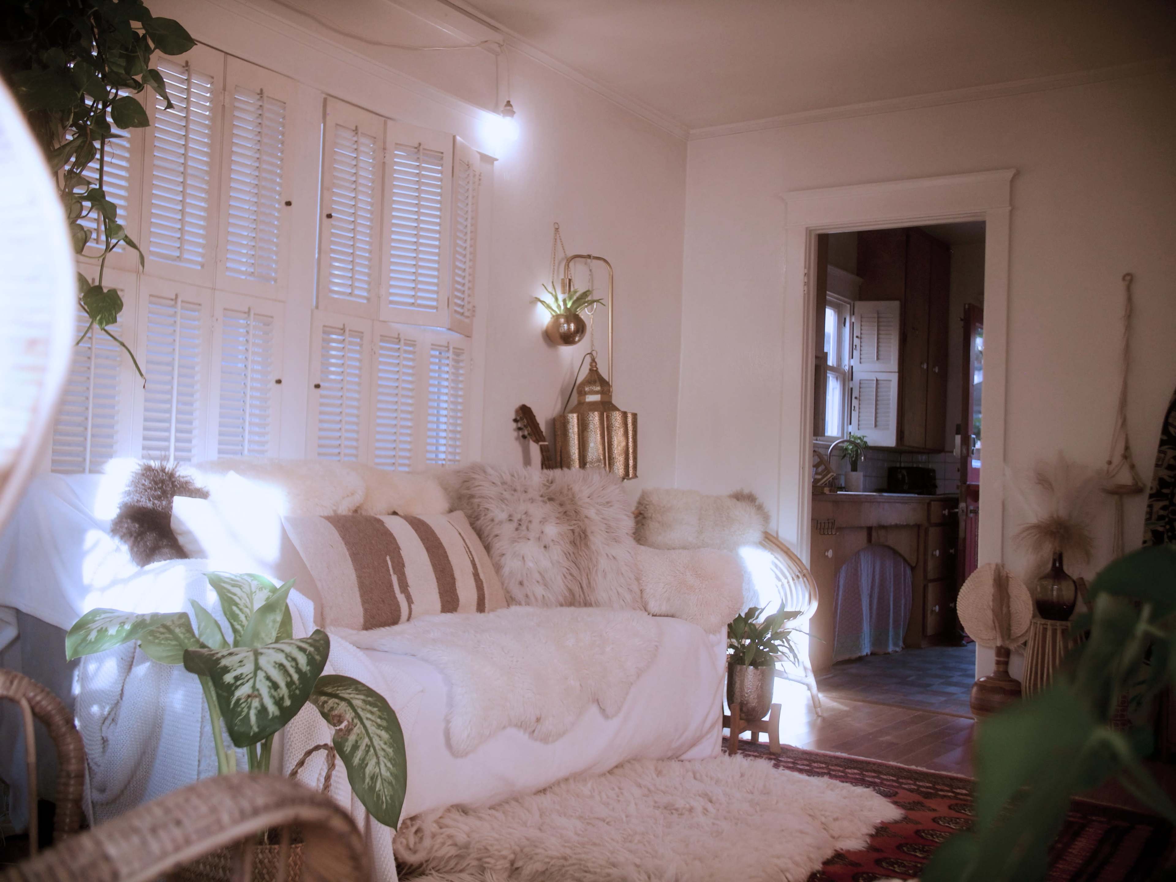 The image shows a cozy living room with a white sofa covered in fur throws, surrounded by potted plants and a doorway leading to a kitchen.