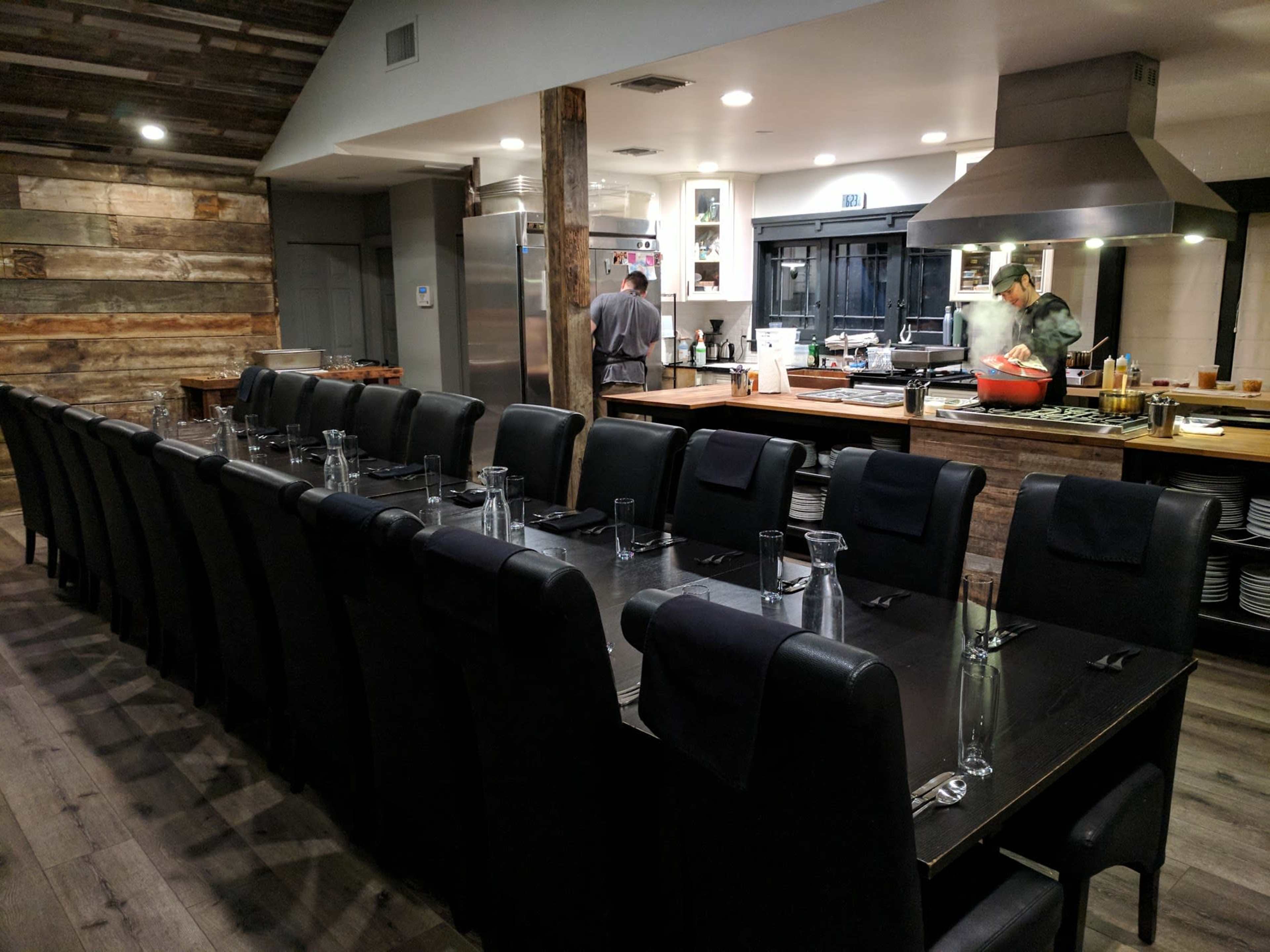 The image shows a modern restaurant interior with a long, empty dining table and an open kitchen area where two chefs are preparing food.