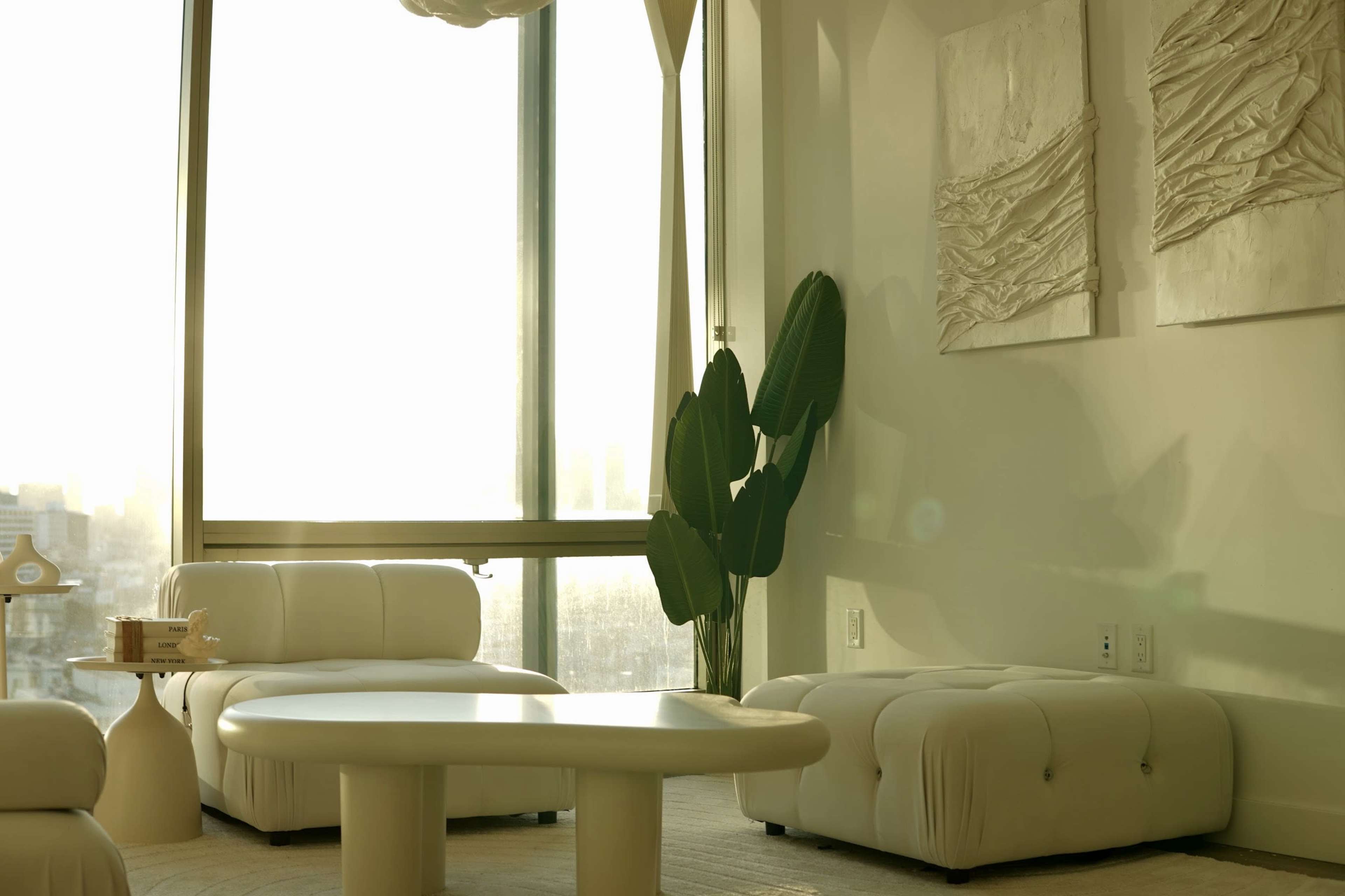 A bright, modern living room features white furniture, a large window, and a green plant beside abstract wall art.