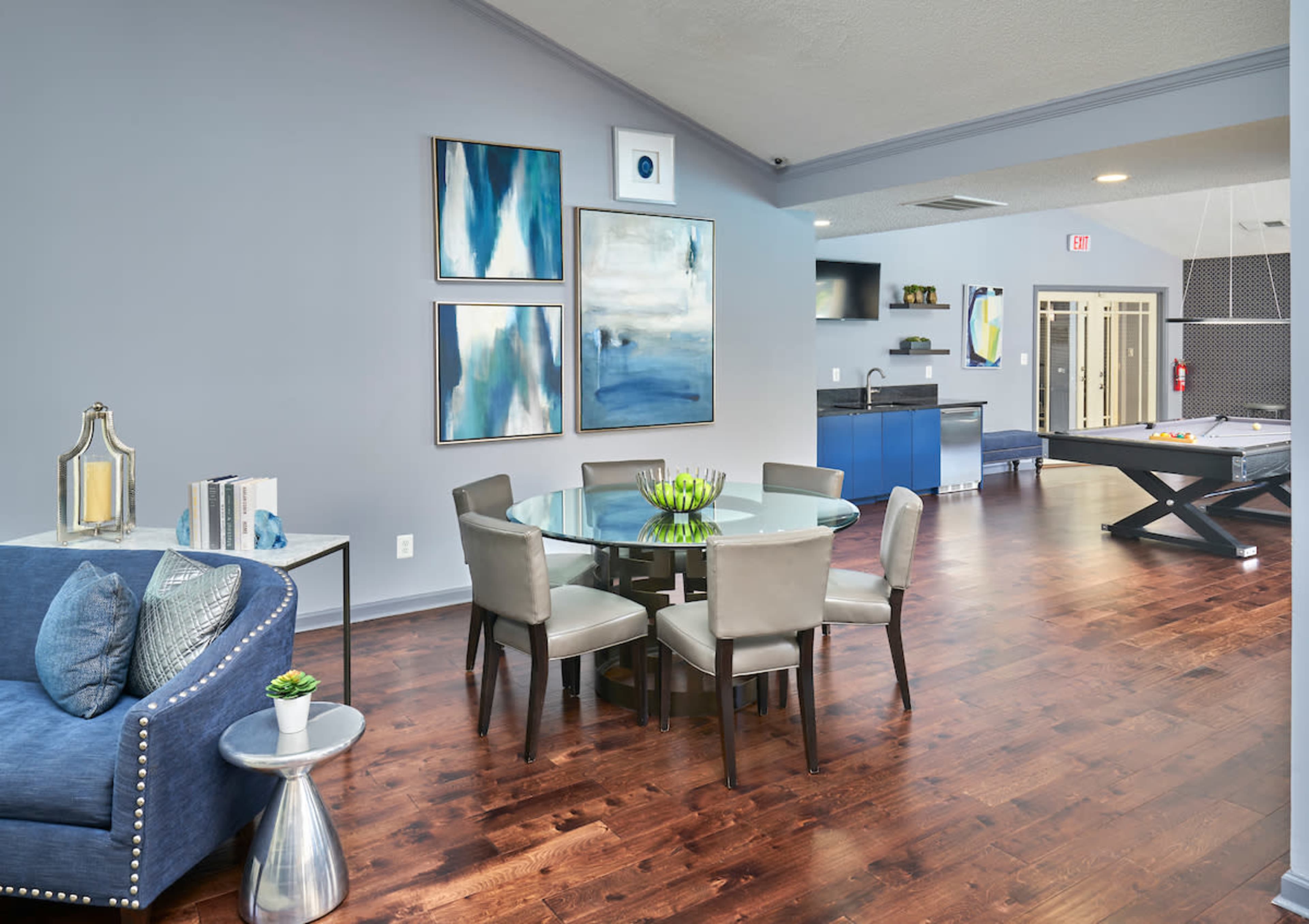 A modern lounge area featuring a round glass dining table with gray chairs, a blue sofa with decorative detailing, and abstract artwork on the walls, all set against a hardwood floor.