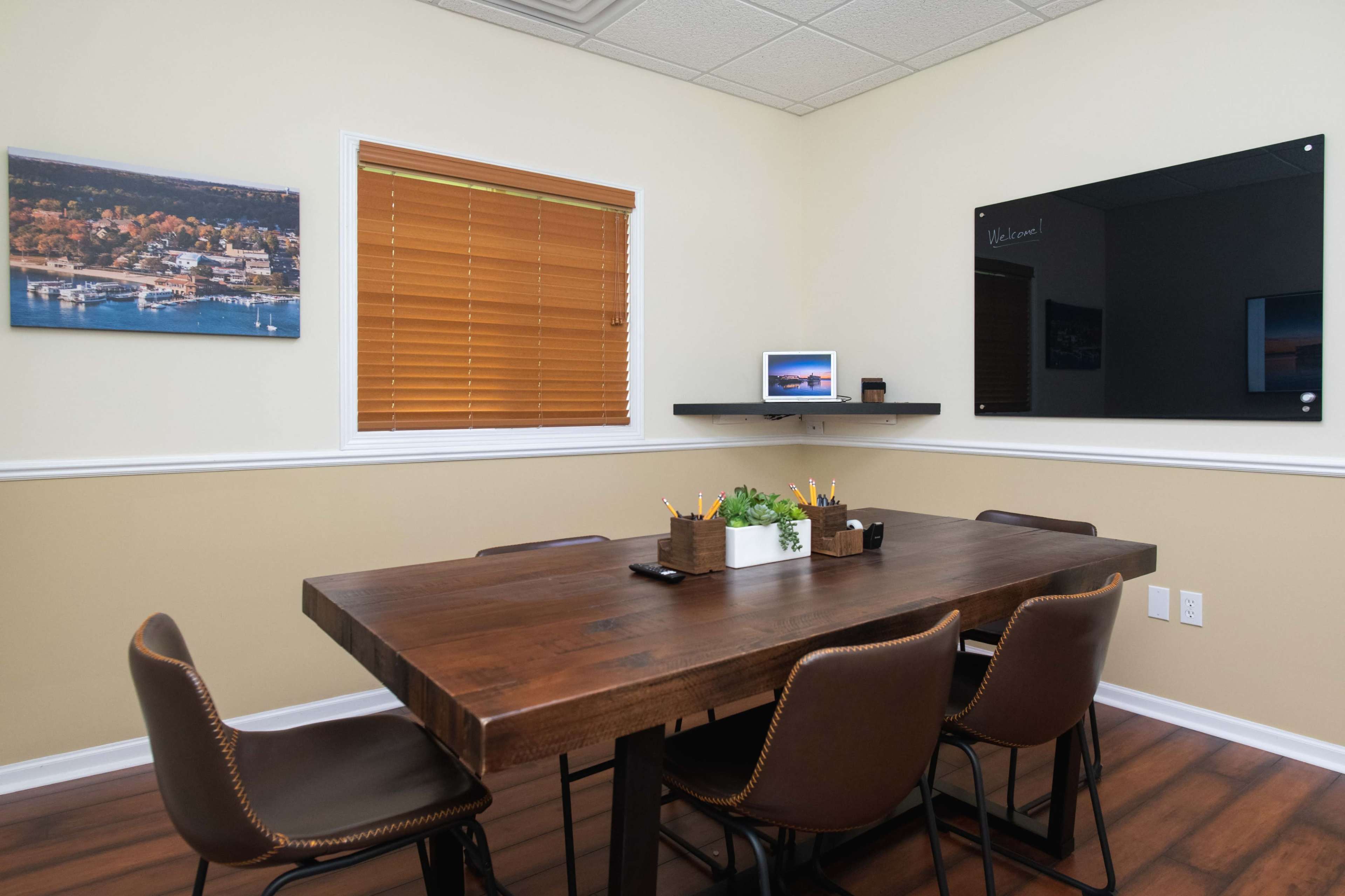 A meeting room features a wooden table surrounded by leather chairs, with a plant centerpiece and a wall-mounted shelf displaying a small screen and a photo.