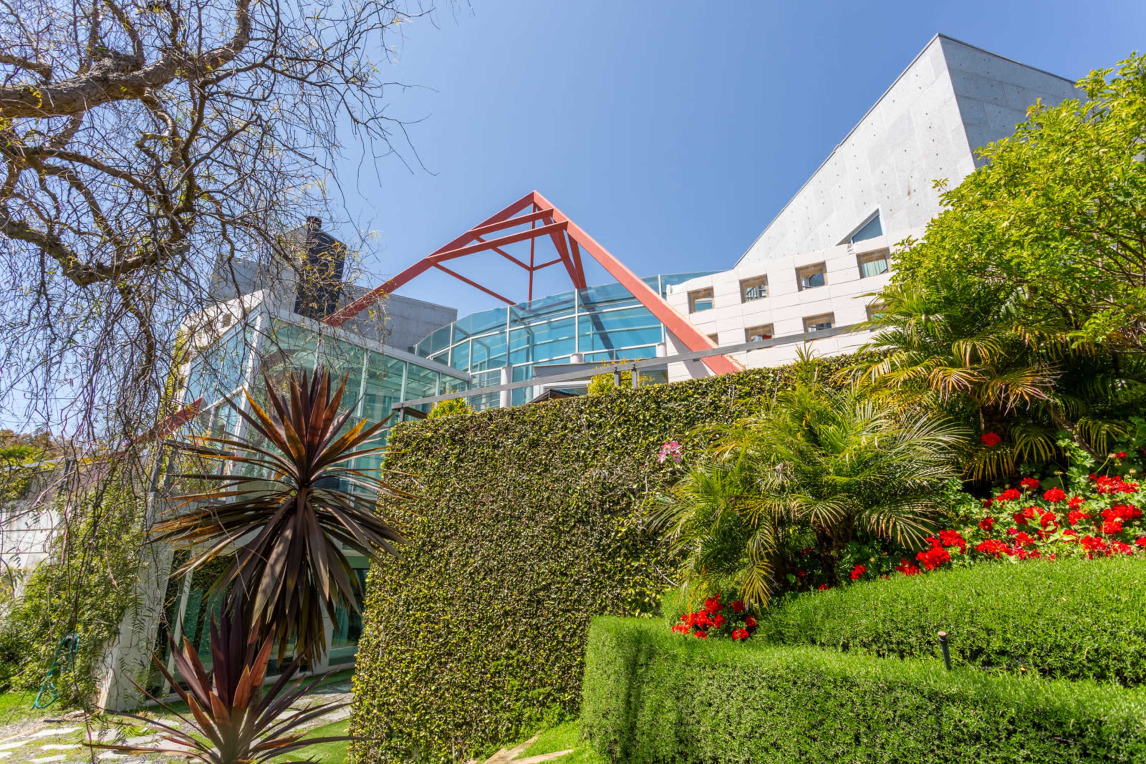 A modern architectural building featuring glass elements and a prominent red triangular roof, surrounded by greenery and vibrant flowers.