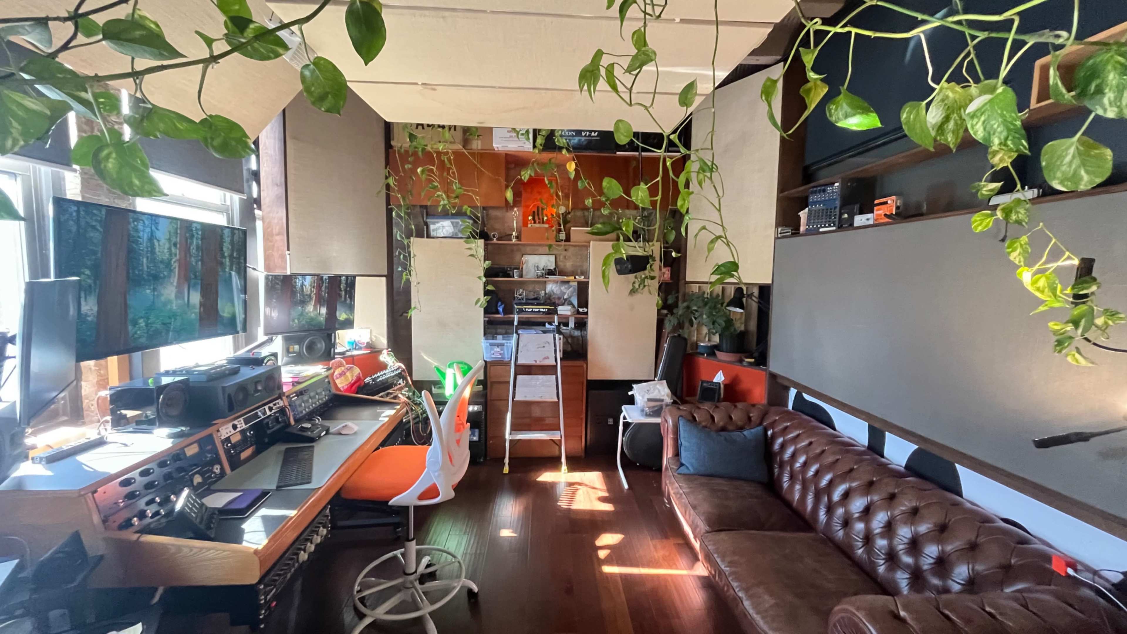 The image shows a cozy office space featuring a brown leather sofa, a desk with audio equipment and computer monitors, and several hanging plants.