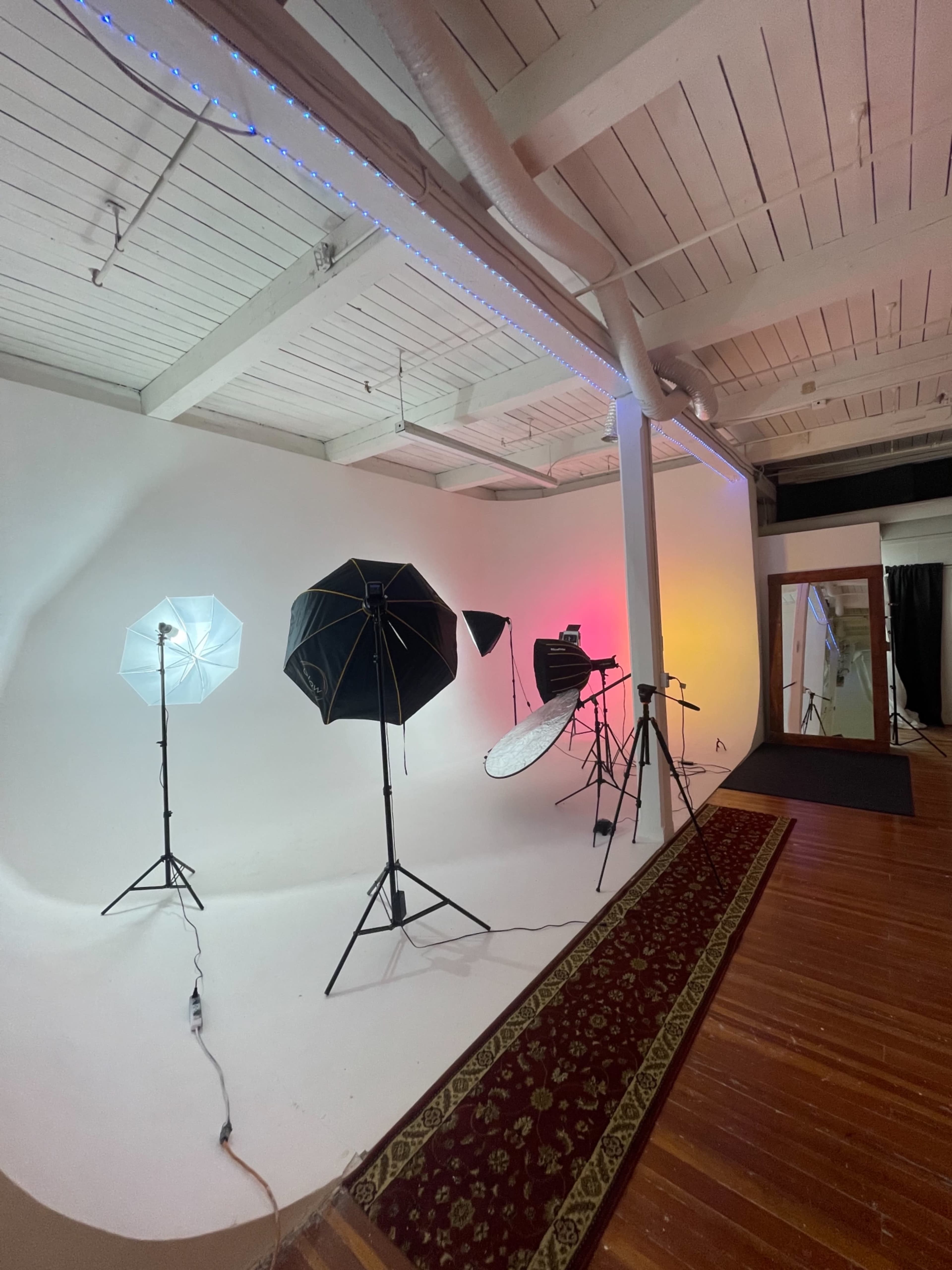 The image shows a photography studio with several light umbrellas and a large mirror set against a white backdrop and wooden floor.