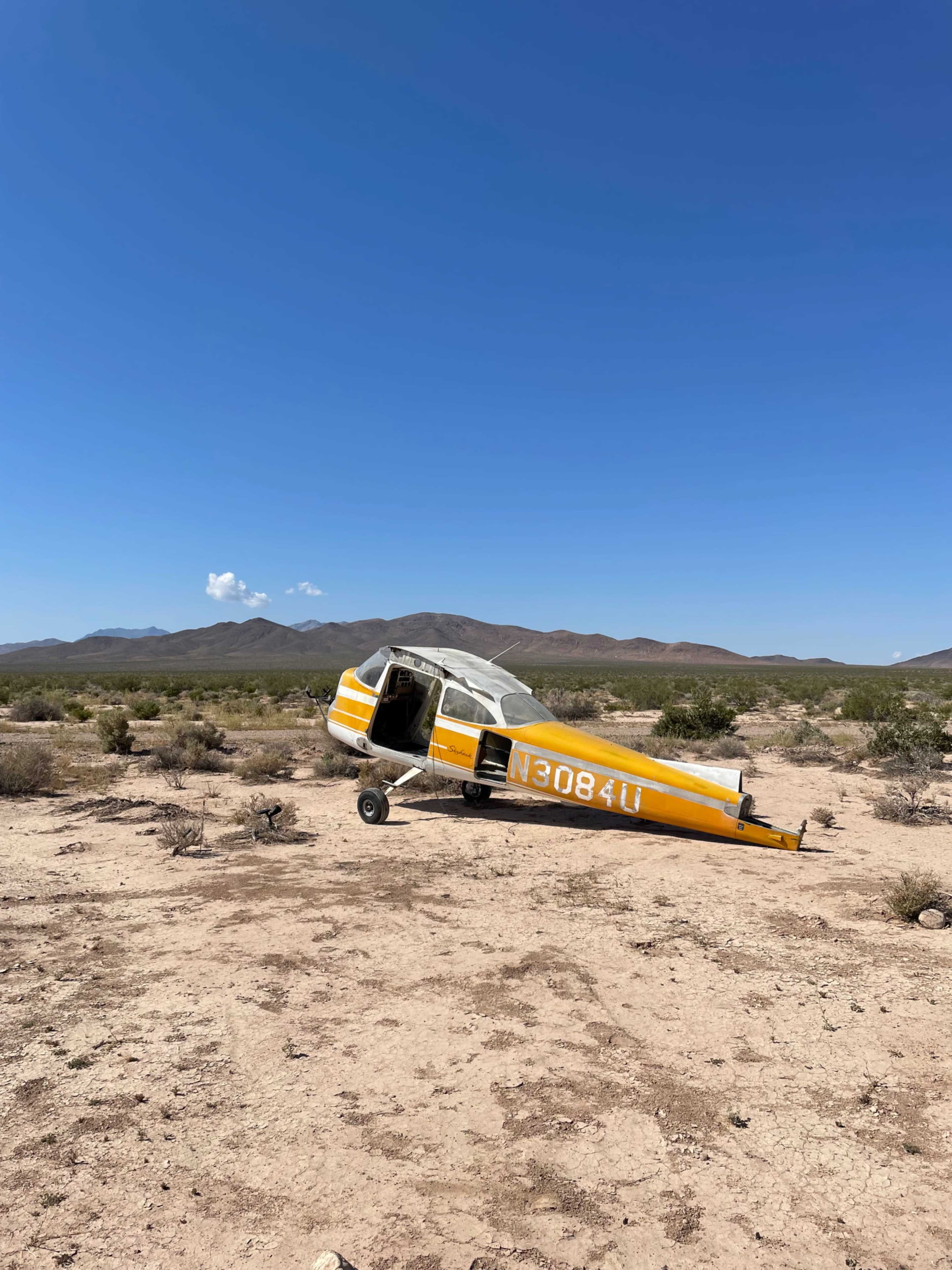 An abandoned yellow and white aircraft is partially upright in a dry desert landscape with distant mountains under a clear blue sky.