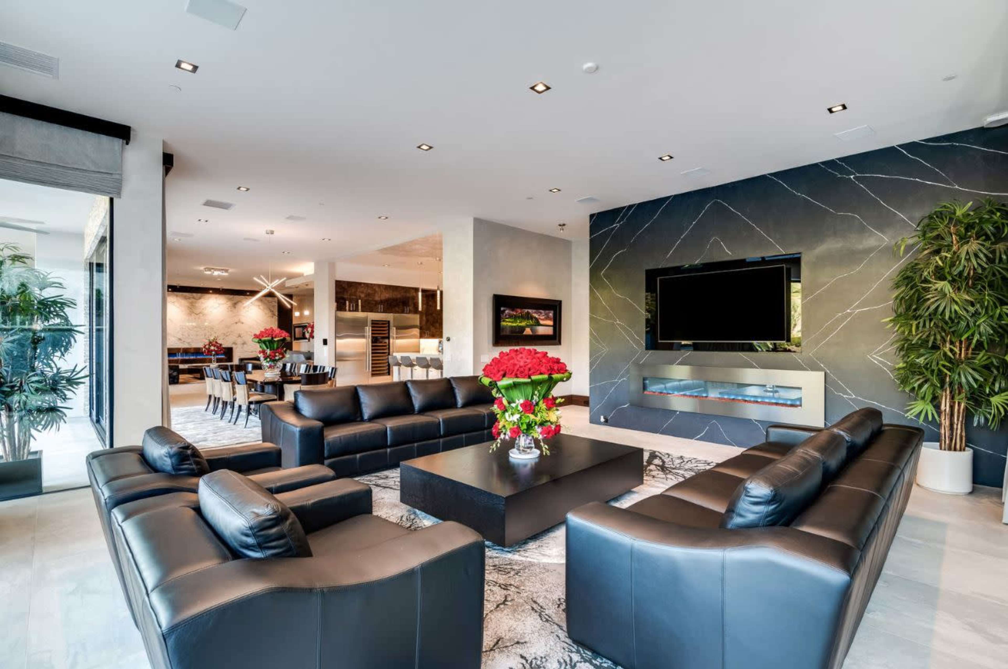 A modern living room features black leather sofas arranged around a central coffee table, with a large TV mounted on a marble wall and greenery in the corners.