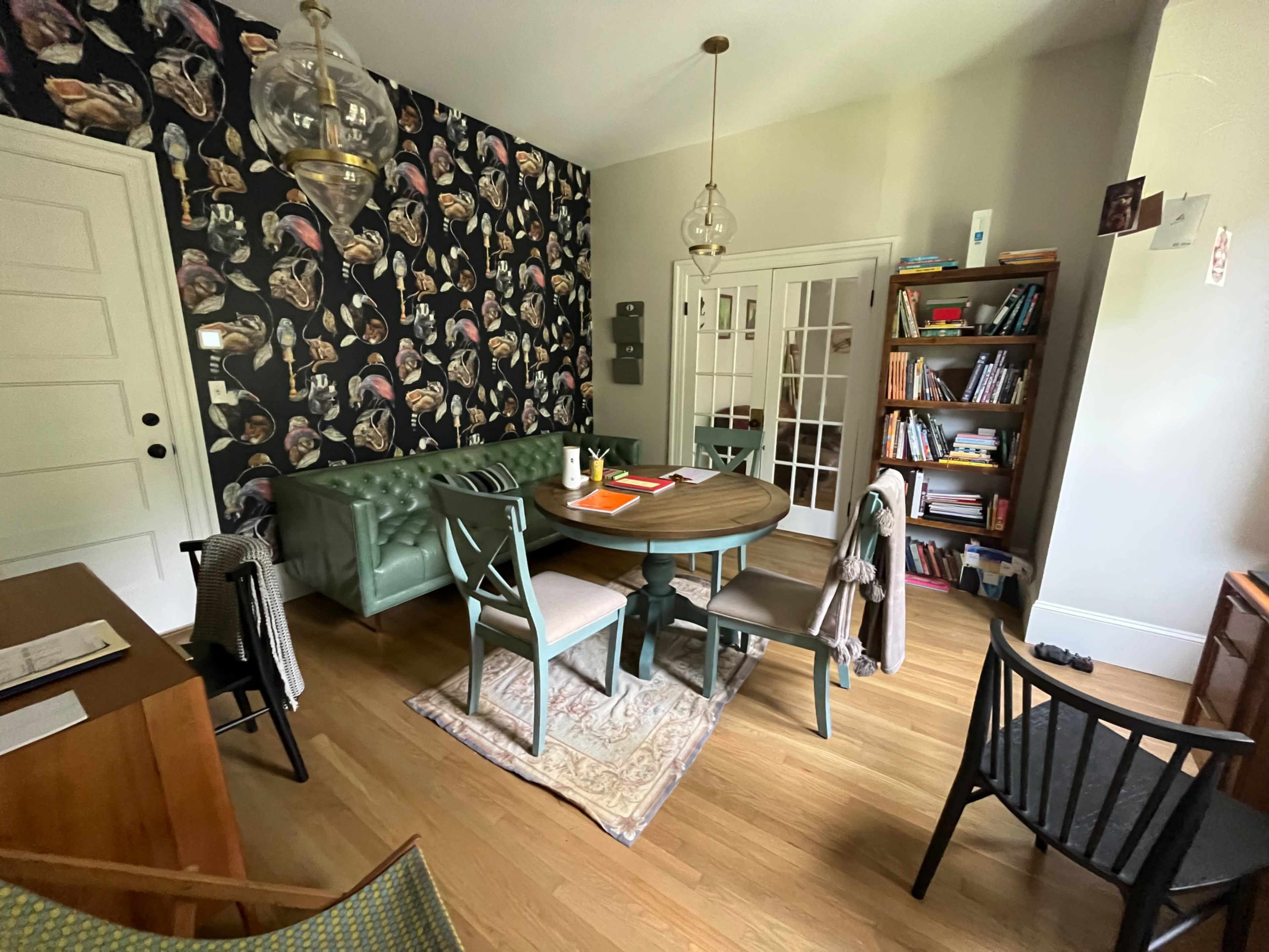 A cozy room featuring a round wooden dining table surrounded by chairs, a patterned wallpaper, a green upholstered bench, and a bookshelf filled with books.