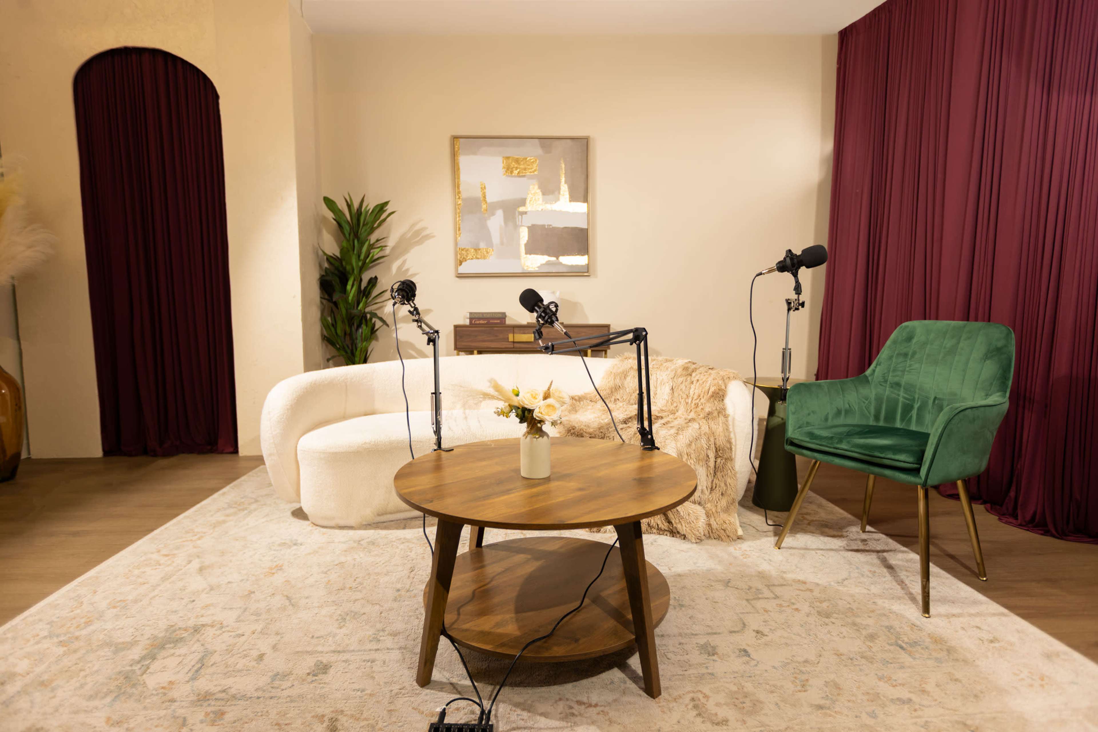 The image shows a podcast or interview setup featuring a light-colored sofa, a green chair, a round wooden coffee table with a small vase of flowers, and two microphones positioned on stands.