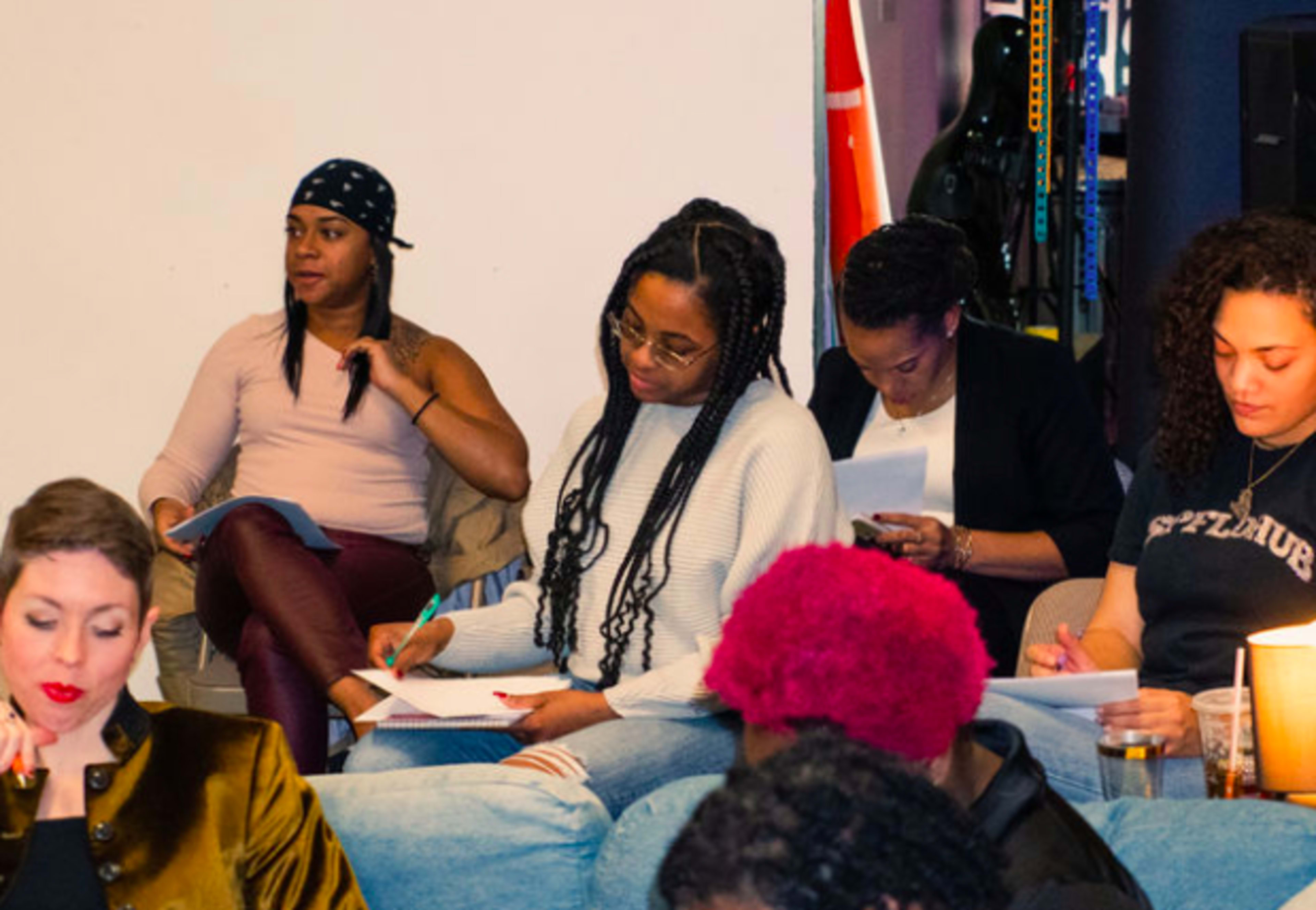 A group of four women are sitting in a casual setting, writing notes and engaging with a discussion.