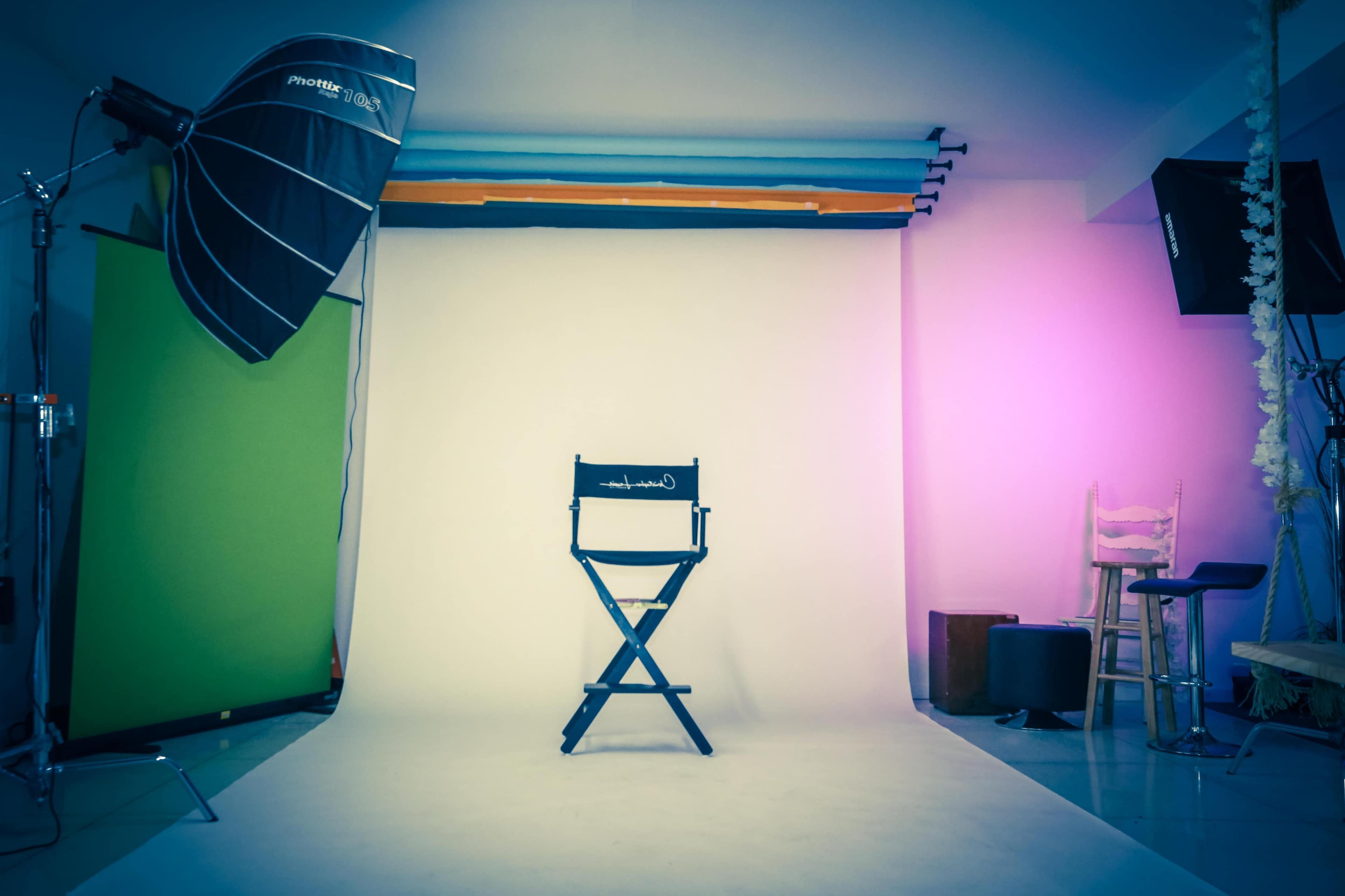 A photography studio is set up with a director's chair in front of a blank backdrop, surrounded by lighting equipment and colored curtains.
