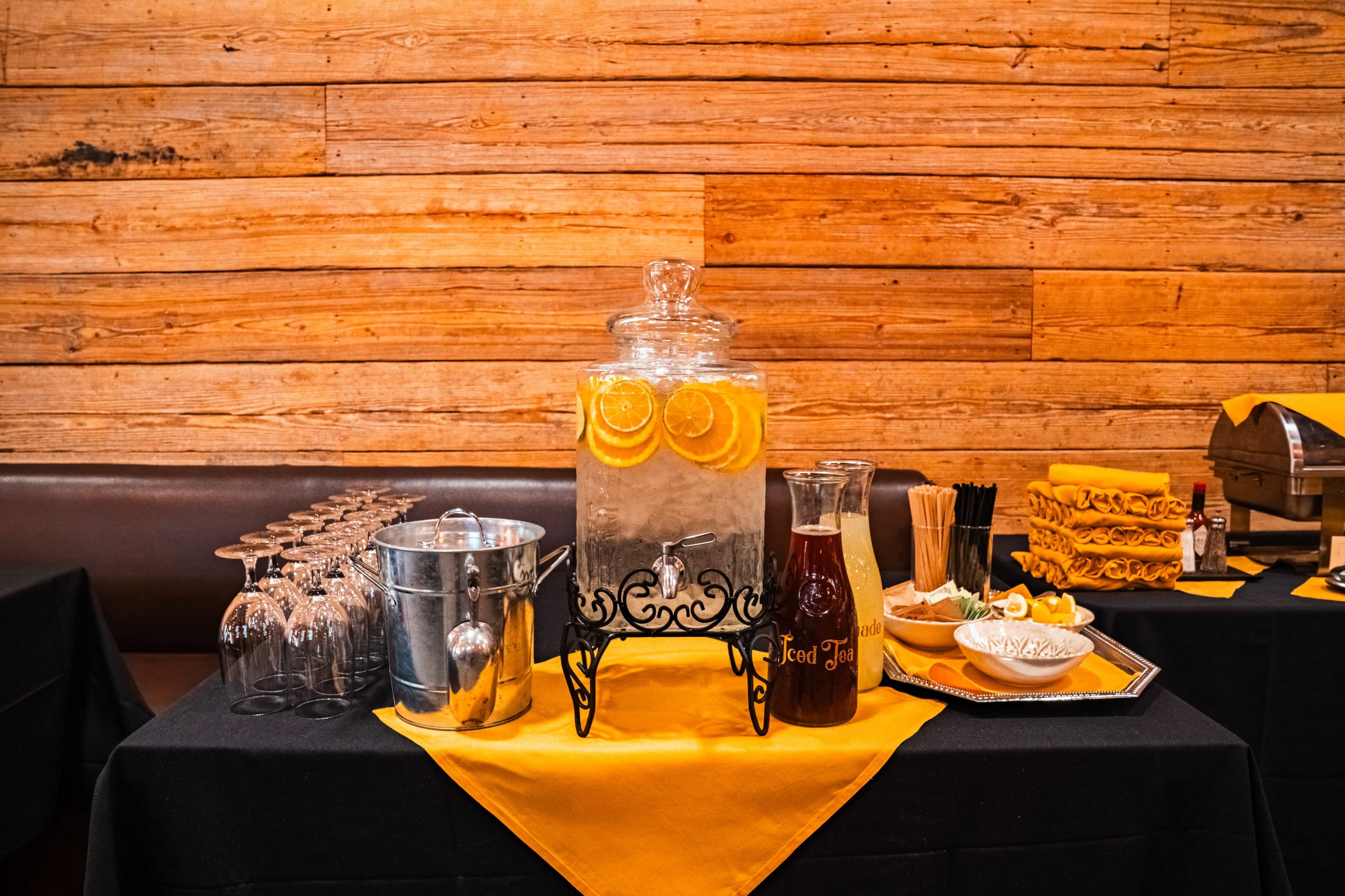 A beverage station features a glass dispenser filled with water and orange slices, accompanied by a pitcher of tea, a bucket of ice, glasses, and a tray of garnishes on a black tablecloth against a wooden backdrop.