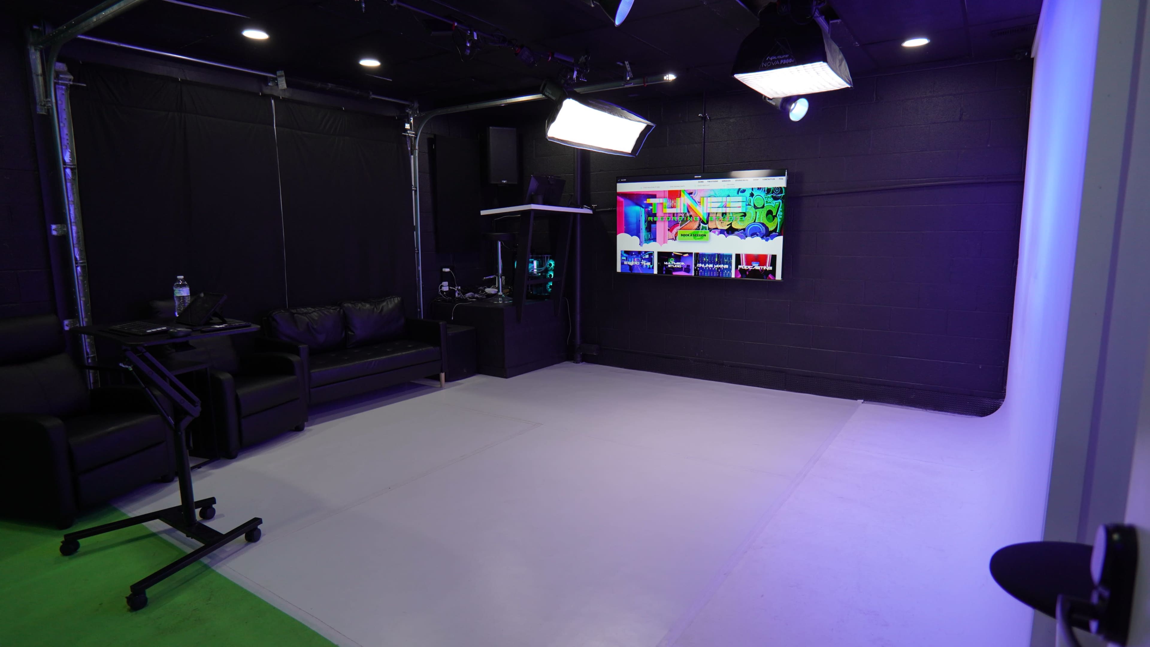 The image shows a small studio space with a black couch, a monitor displaying colorful graphics, and studio lighting set up against a backdrop of black and white walls.